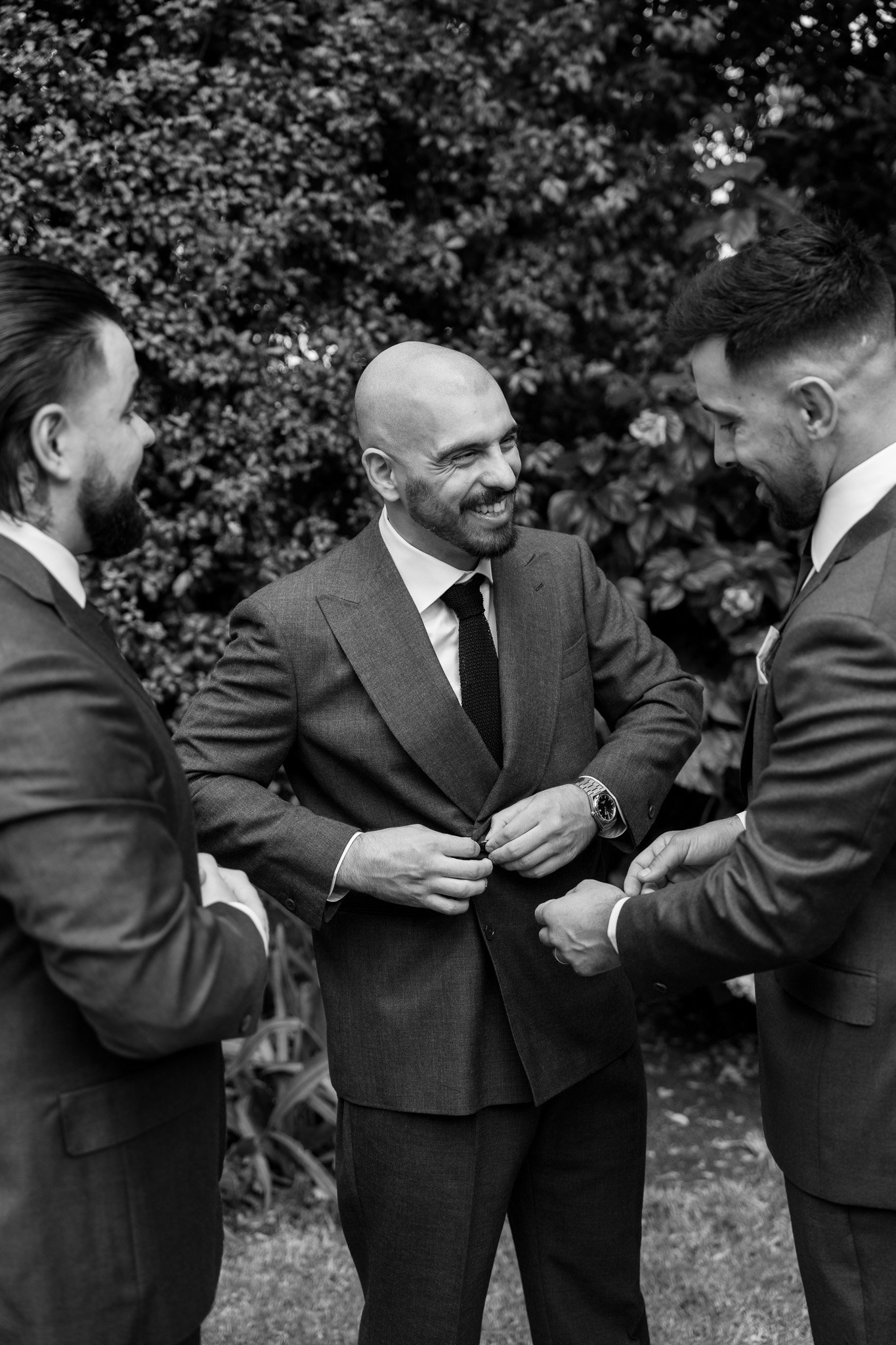 Three men dressed in suits having a conversation outdoors, with a leafy background.