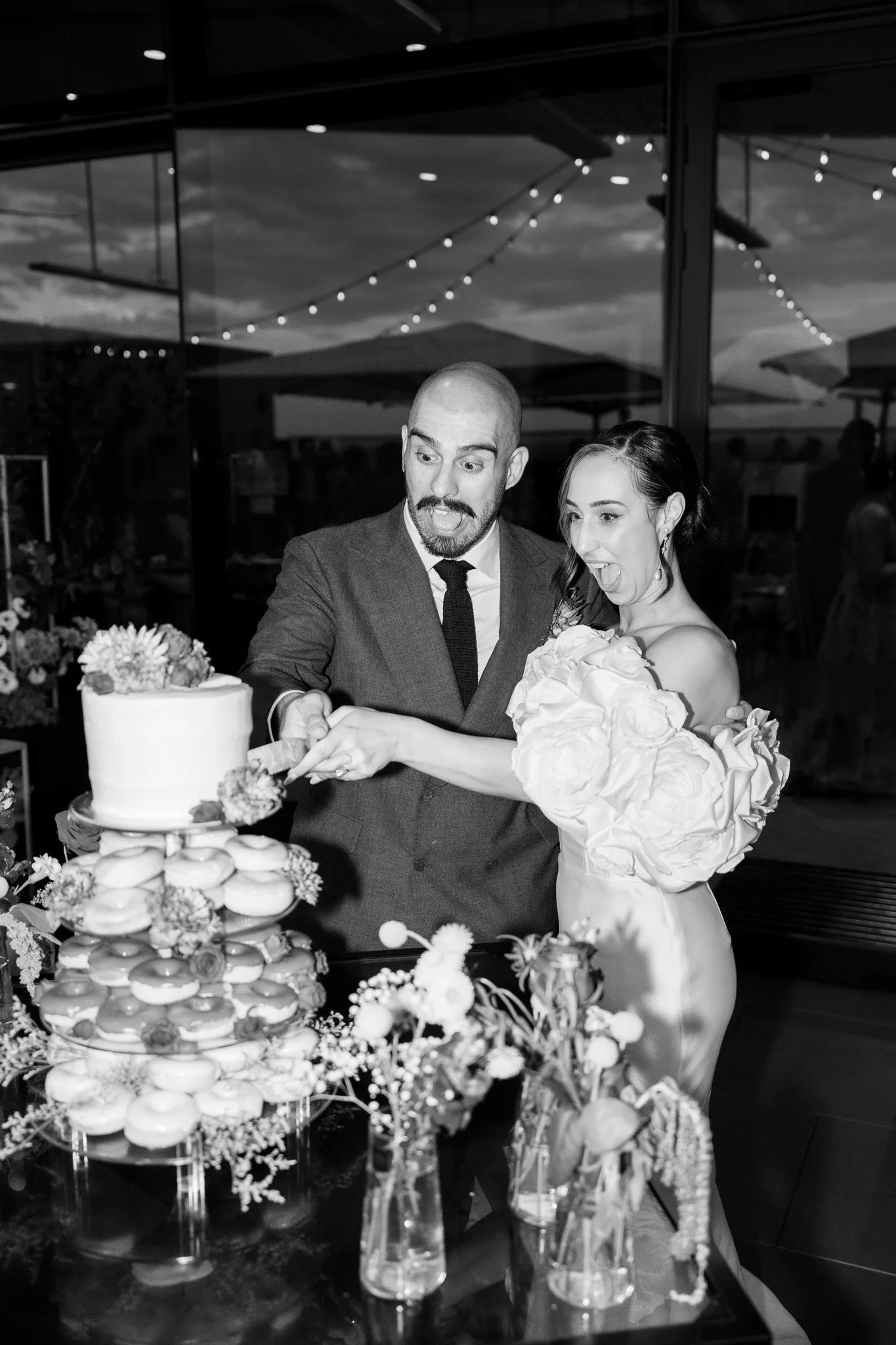 Black-and-white photo of a man and woman cutting a wedding cake at a celebration, with floral decorations and a glass window reflecting string lights.