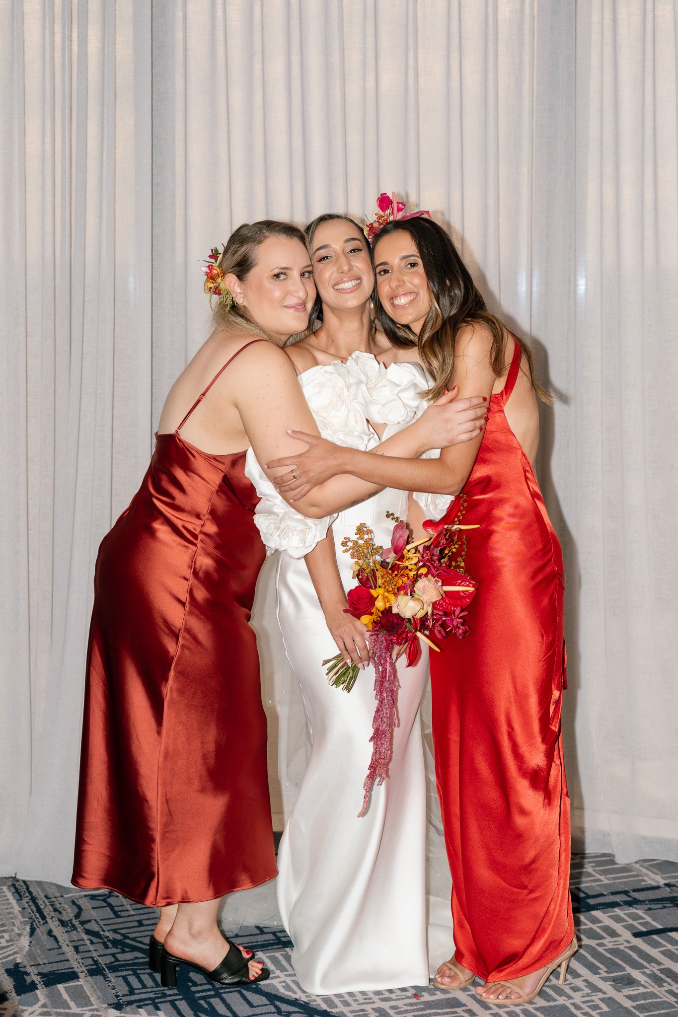 Three women hugging, one in a white wedding dress holding a bouquet, flanked by two women in red satin dresses, standing in front of a white curtain at a wedding.
