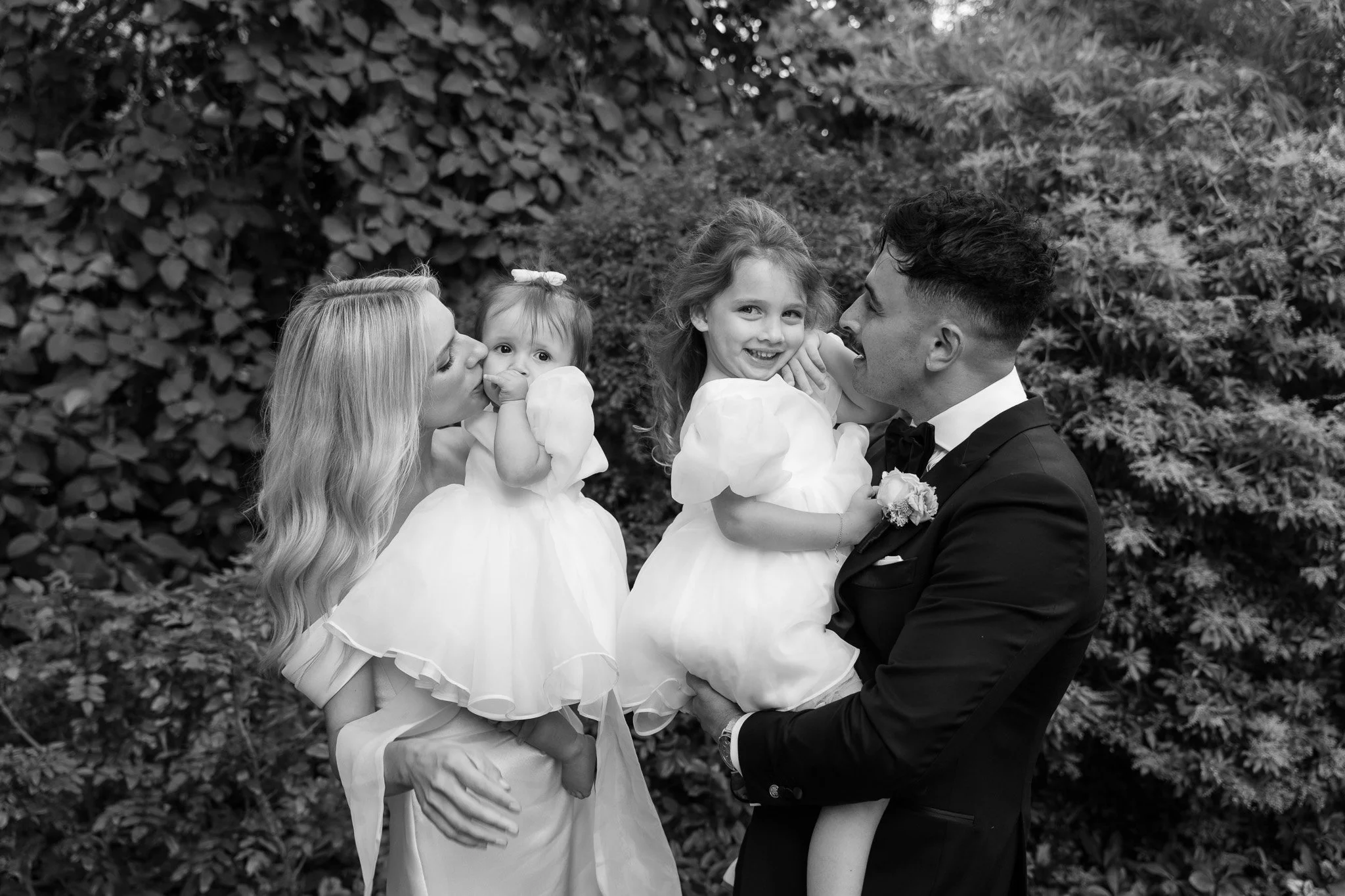 A black and white photo of a happy family at a wedding. The woman is kissing a toddler girl on the cheek while the man in a tuxedo holds a young girl on his lap. The background includes lush foliage.