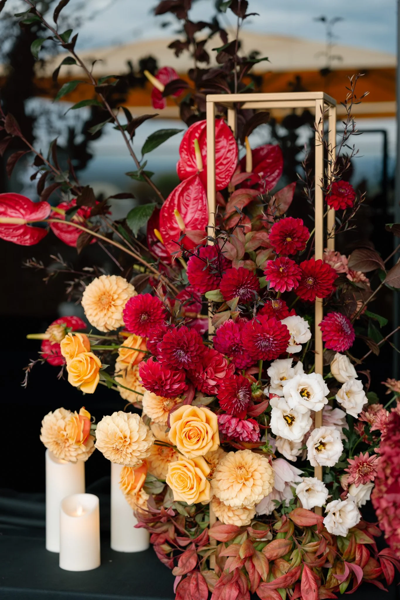Colorful floral arrangement of red, yellow, peach, and white flowers with candles in the foreground and a wooden frame within the arrangement