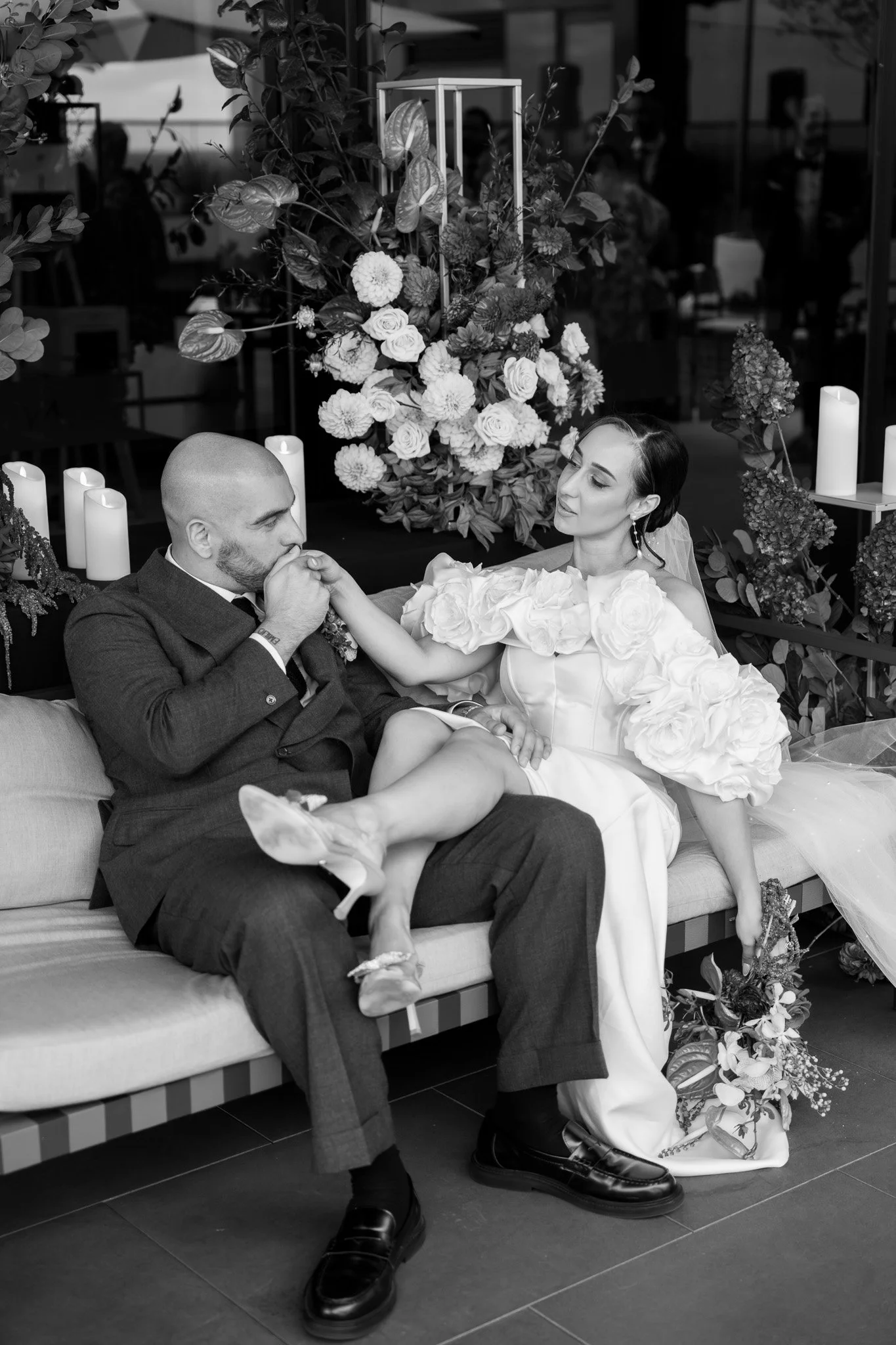 A black-and-white photo of a bride and groom seated on a sofa, with the groom kissing the bride's hand. The bride is wearing a wedding dress with large floral embellishments on the sleeves and holding a bouquet, while the groom is dressed in a suit. 