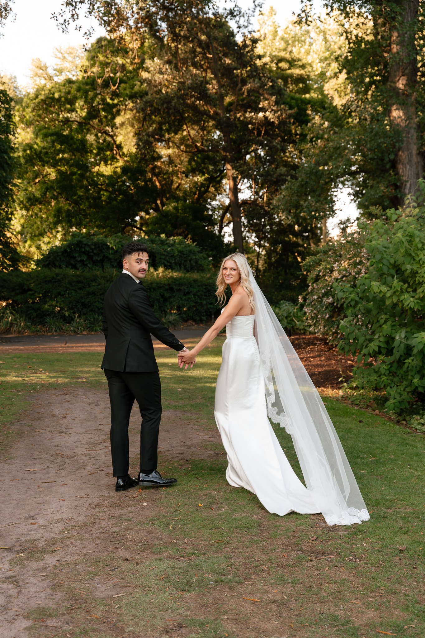 A newly married couple holding hands outdoors on a dirt path in a park surrounded by green trees and bushes, with the bride wearing a white wedding dress and veil, and the groom in a black suit and tie, during late afternoon or early evening with war