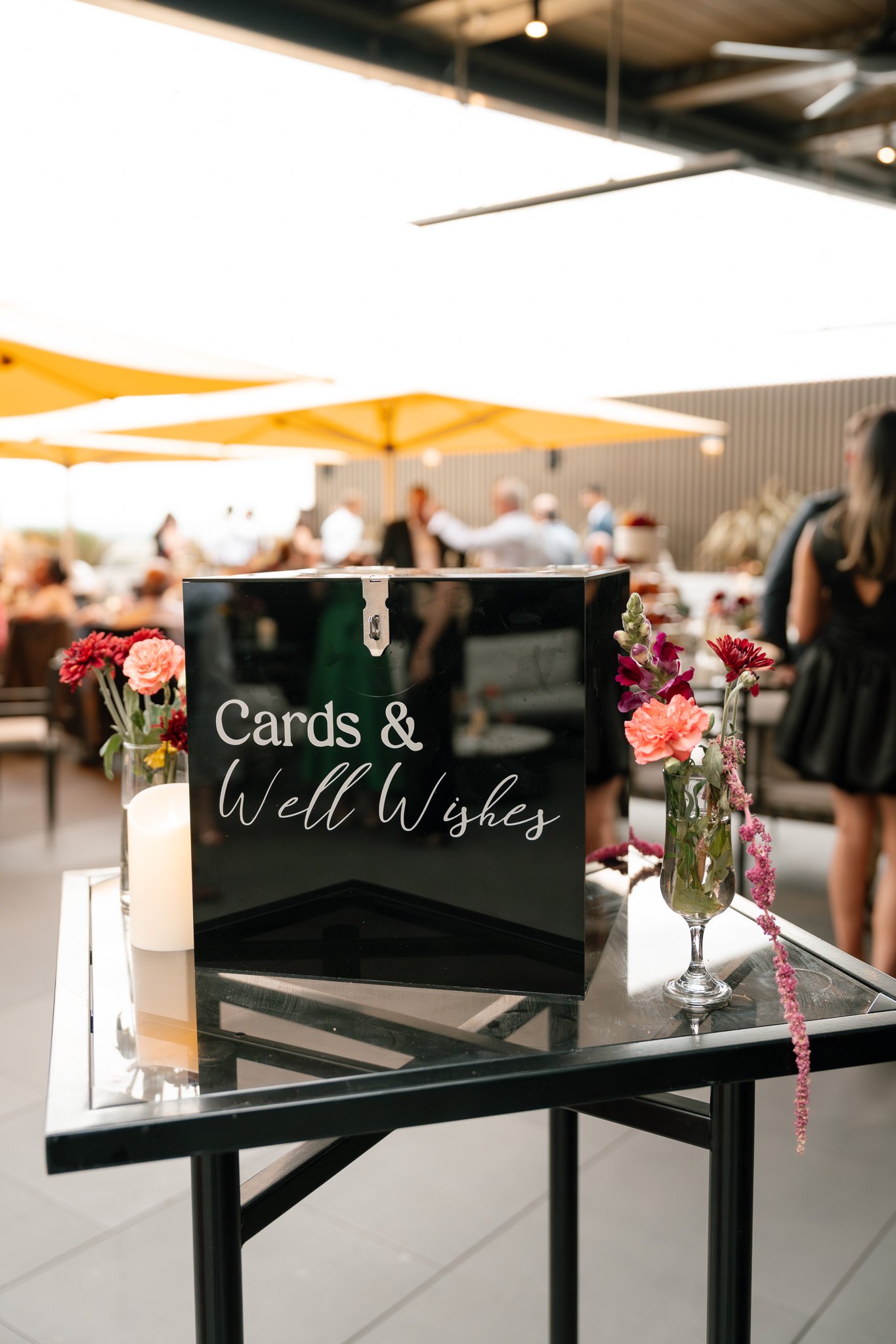 A black sign on a table at a social event reads "Cards & Well Wishes." The table is decorated with pink and red flowers in glass vases, and there are candles nearby. In the background, people are gathered under large yellow umbrellas at an outdoor ve