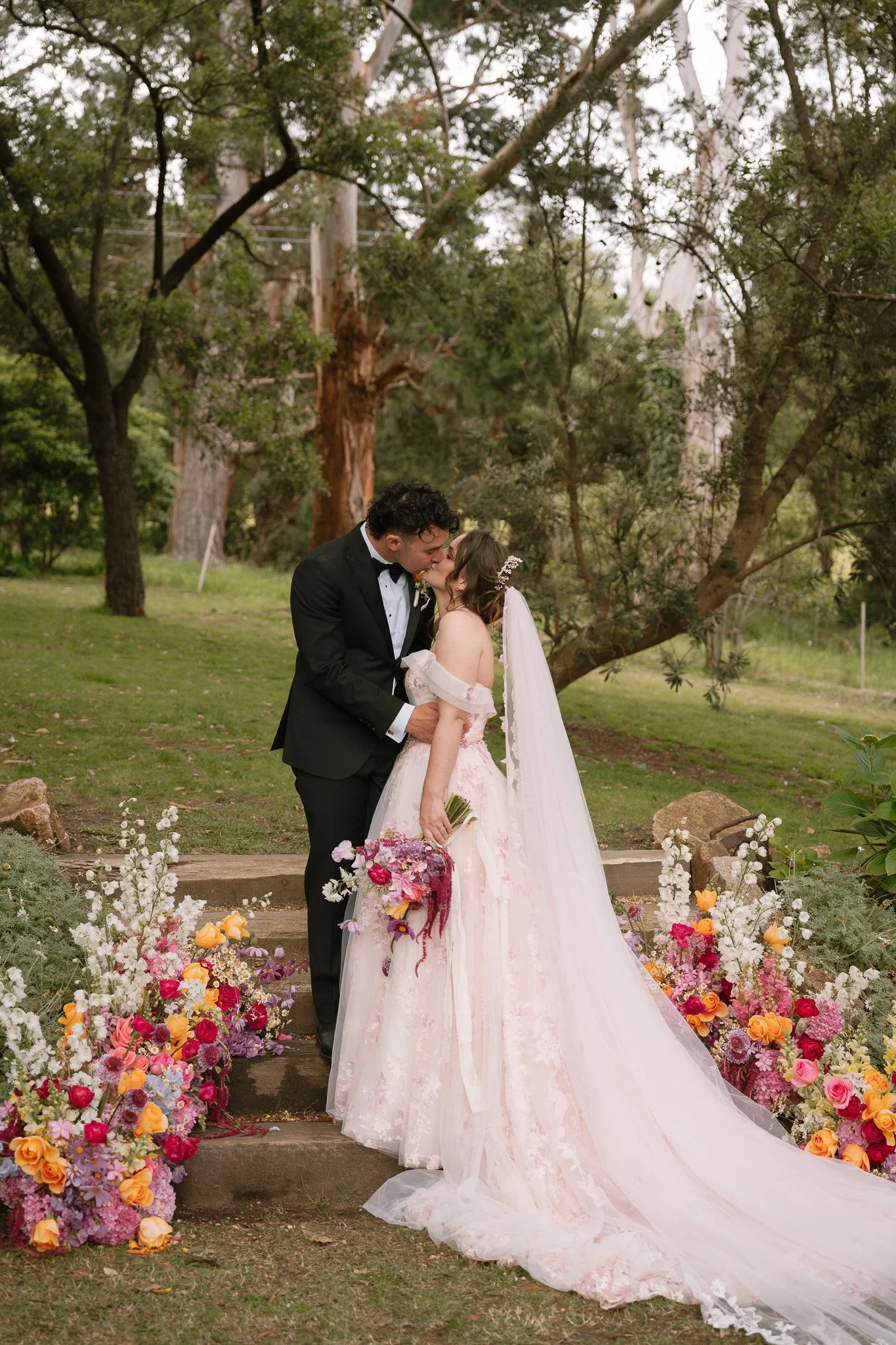 A bride and groom share a kiss outdoors during their wedding, surrounded by colorful flowers and trees.