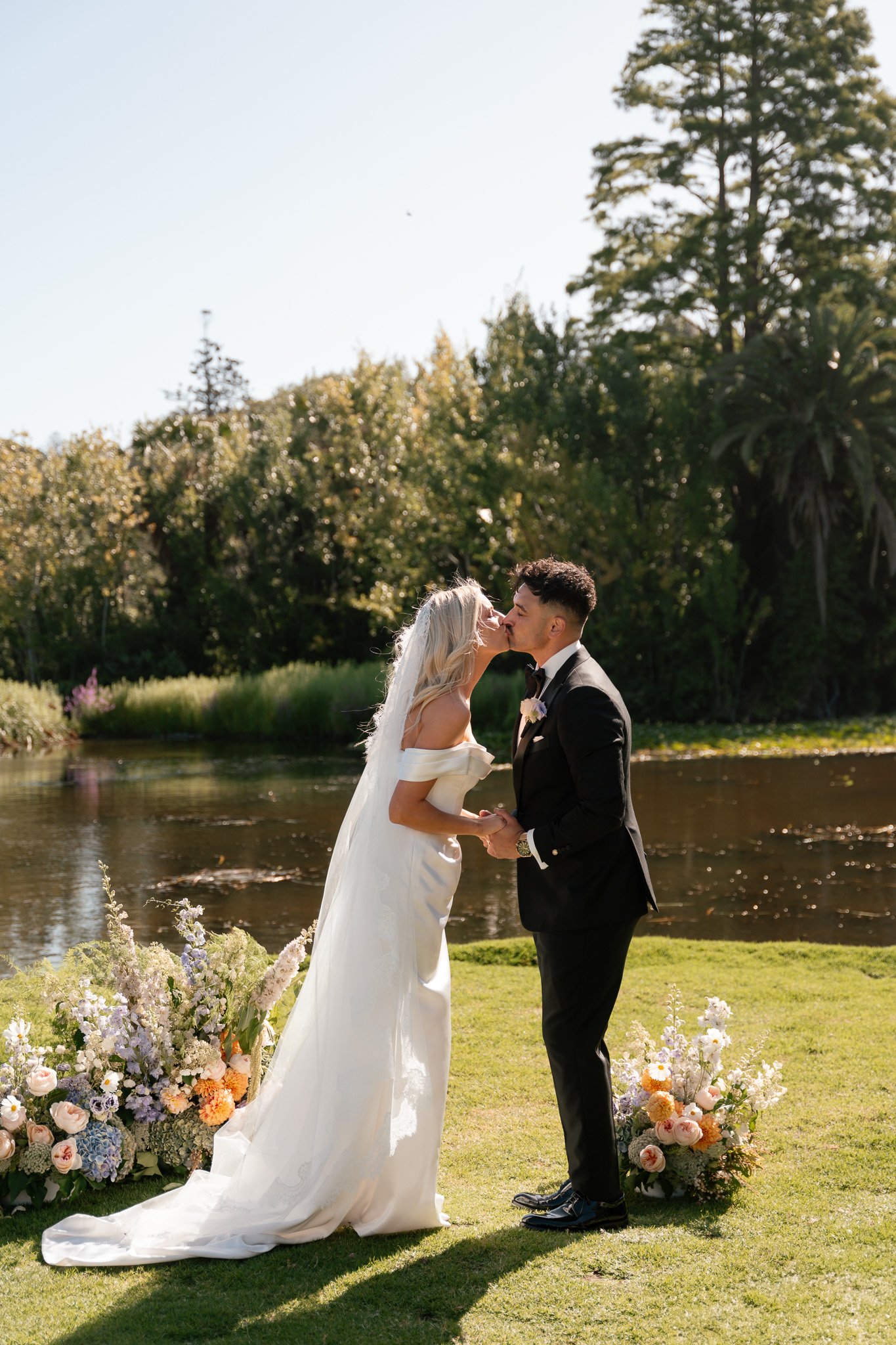 A bride and groom kissing outdoors near a river, with floral arrangements at their feet and trees in the background.
