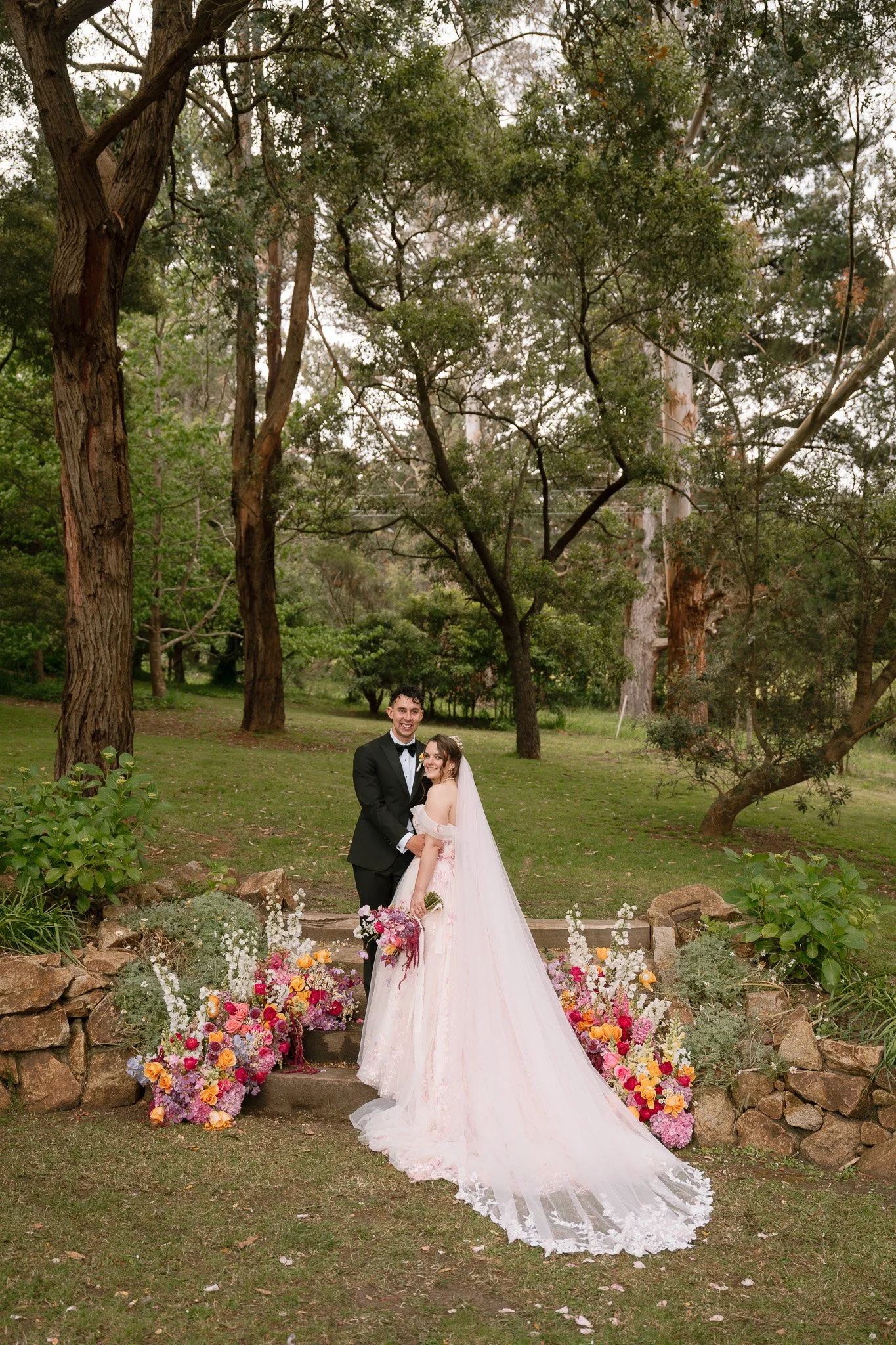 A bride and groom in wedding attire standing on stone steps surrounded by colorful flowers in a lush, green outdoor setting with tall trees.