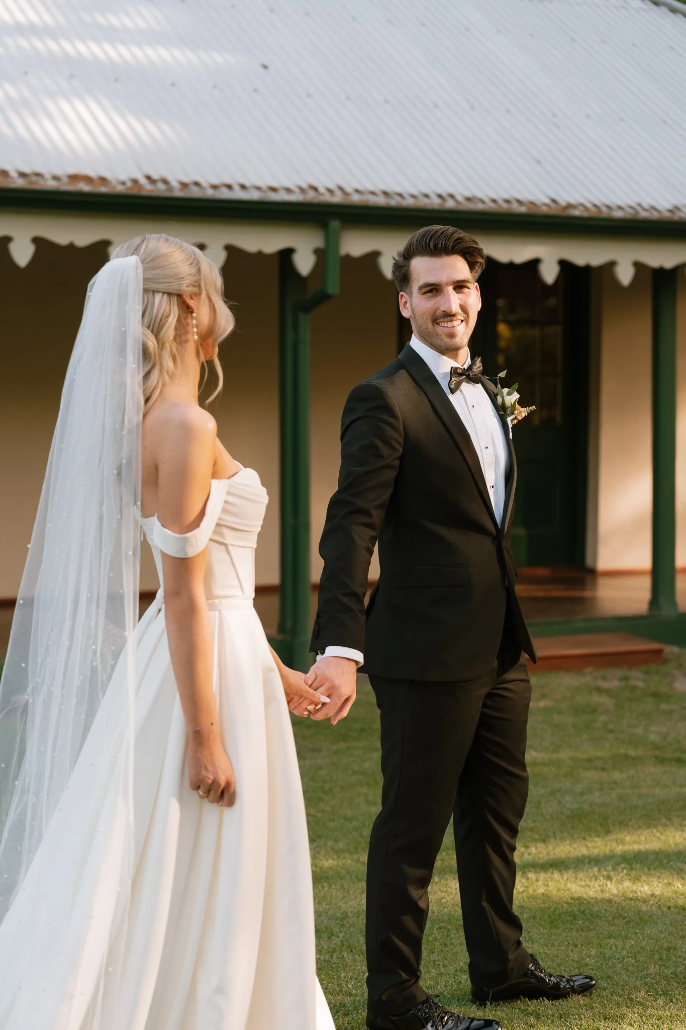 A bride and groom holding hands during their wedding ceremony outdoors, with the groom smiling at the camera and the bride looking towards him, in front of a rustic building with a tin roof.