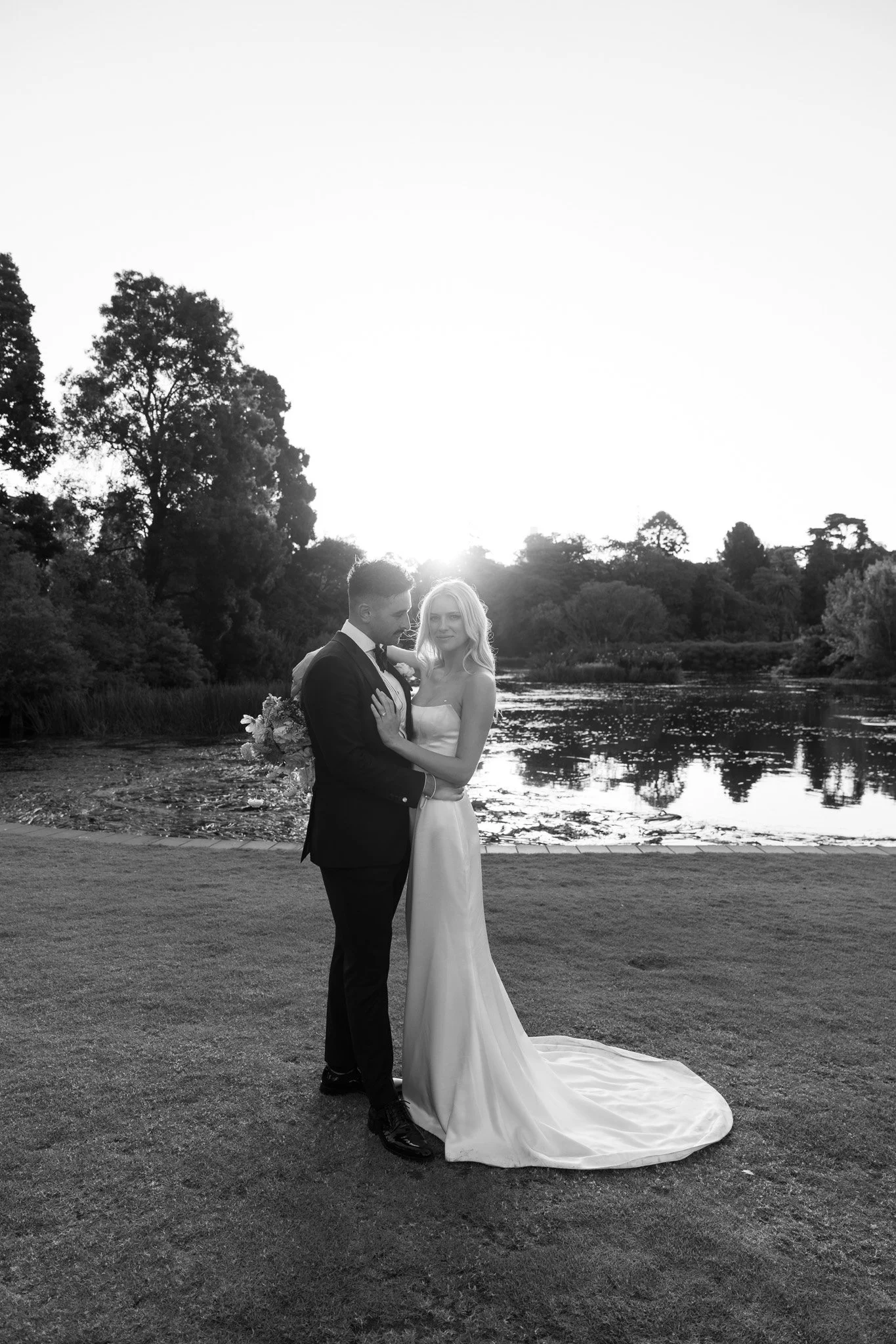 A black and white photo of a bride and groom standing on grass near a pond with trees in the background during sunset. The couple is embracing, with the groom holding a bouquet and the bride in a long wedding dress.