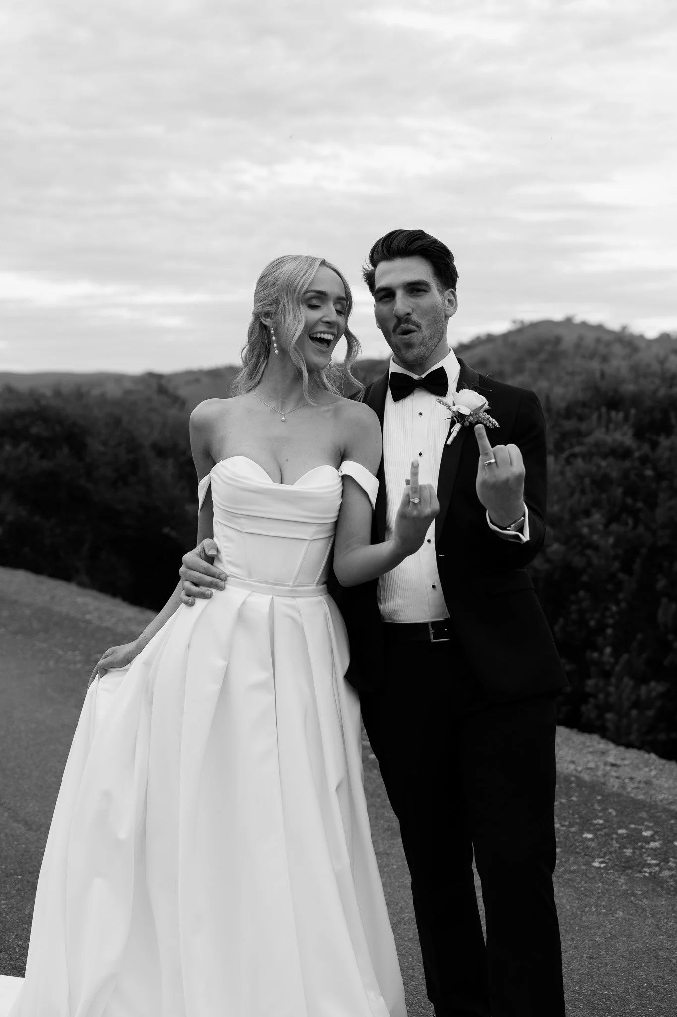 Black-and-white photo of a newlywed couple outdoors, both middle fingers raised, smiling and making playful expressions. The bride wears a strapless wedding gown, the groom a tuxedo with a bow tie.