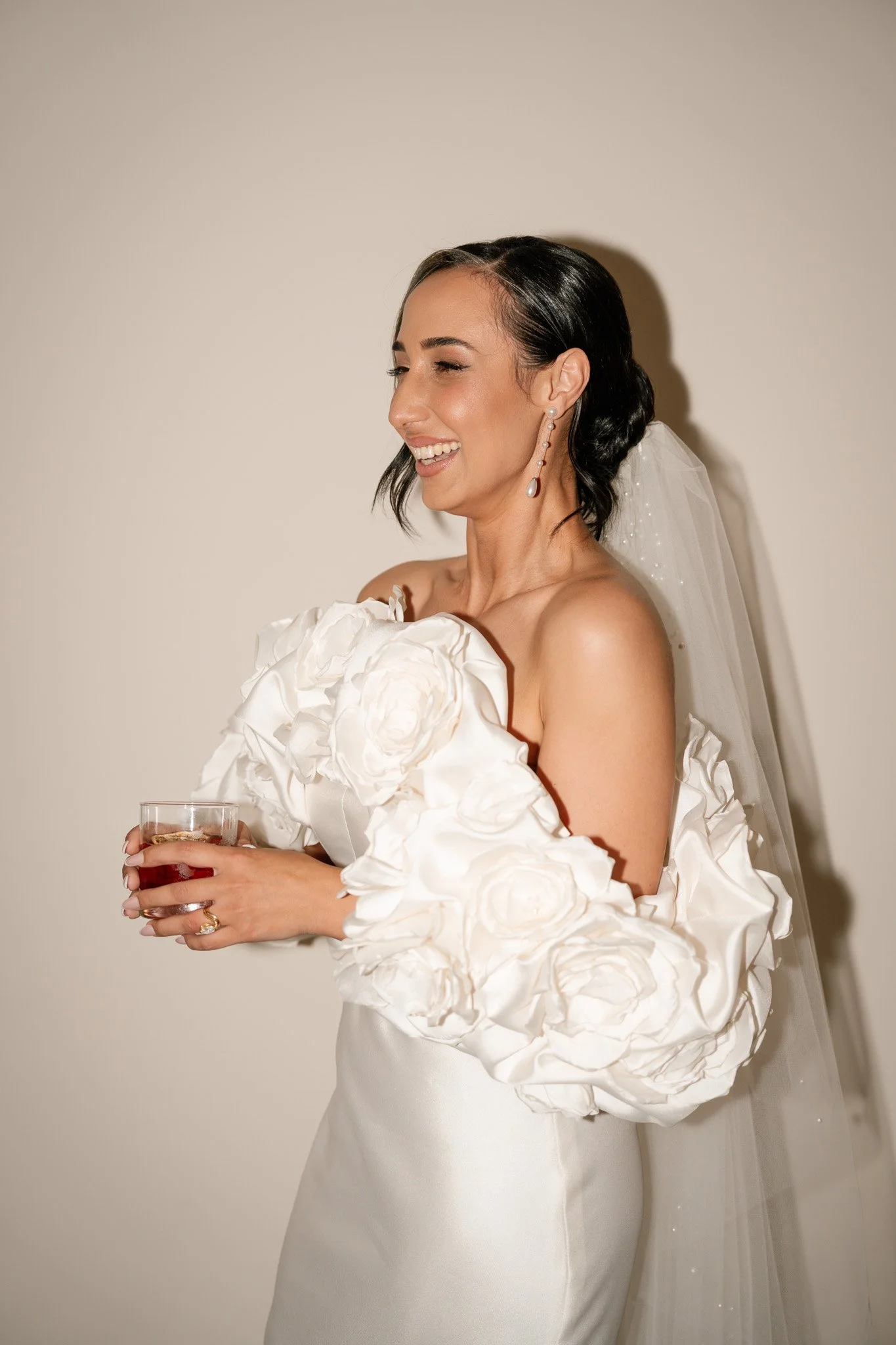 A woman in a white wedding dress with large floral sleeves, holding a glass of drink, smiling, with earrings and a veil.