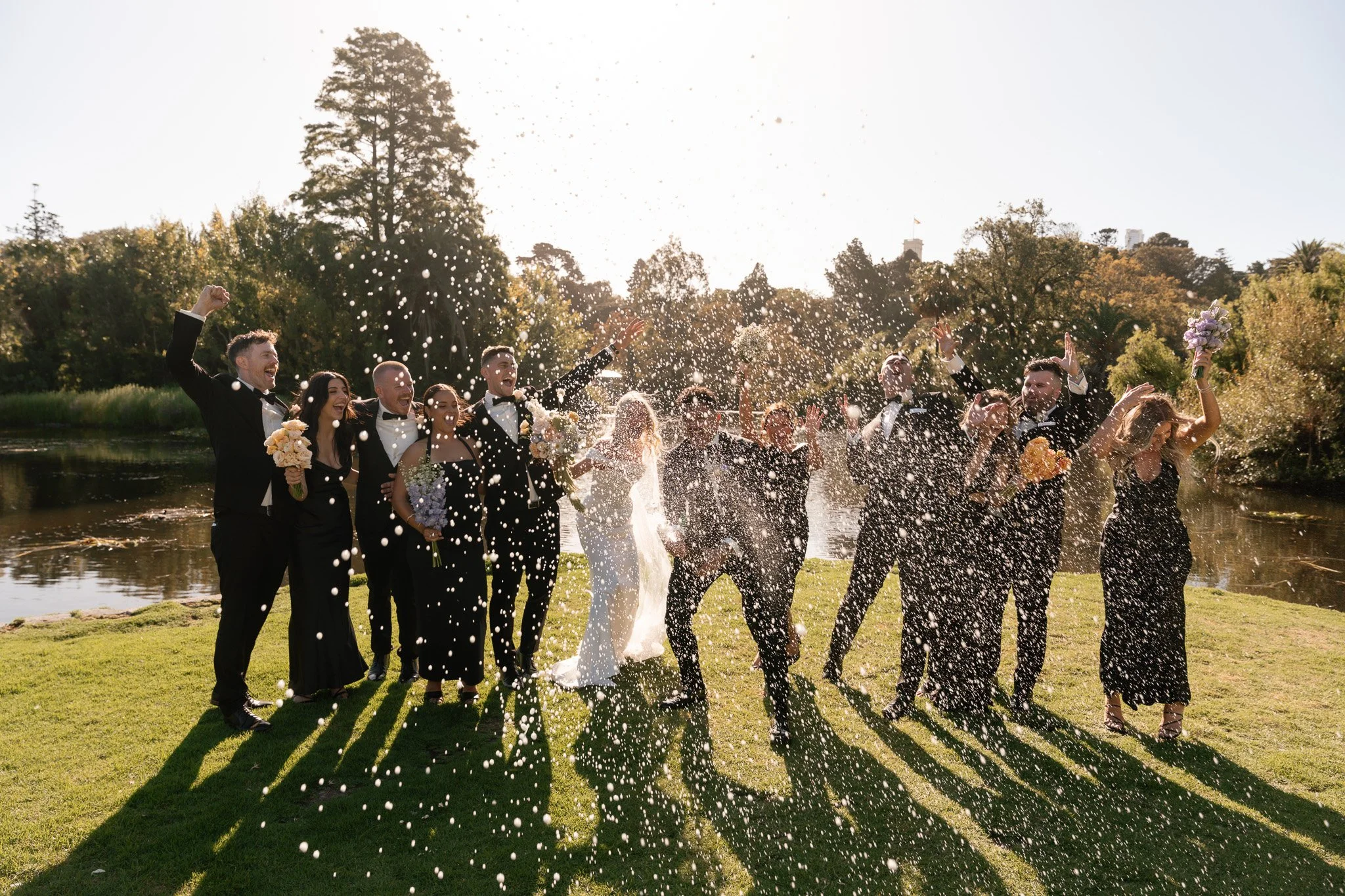 A wedding party celebrating by a river, with guests throwing confetti or rice in the air, dressed in formal suits and dresses, surrounded by trees and sunlight.