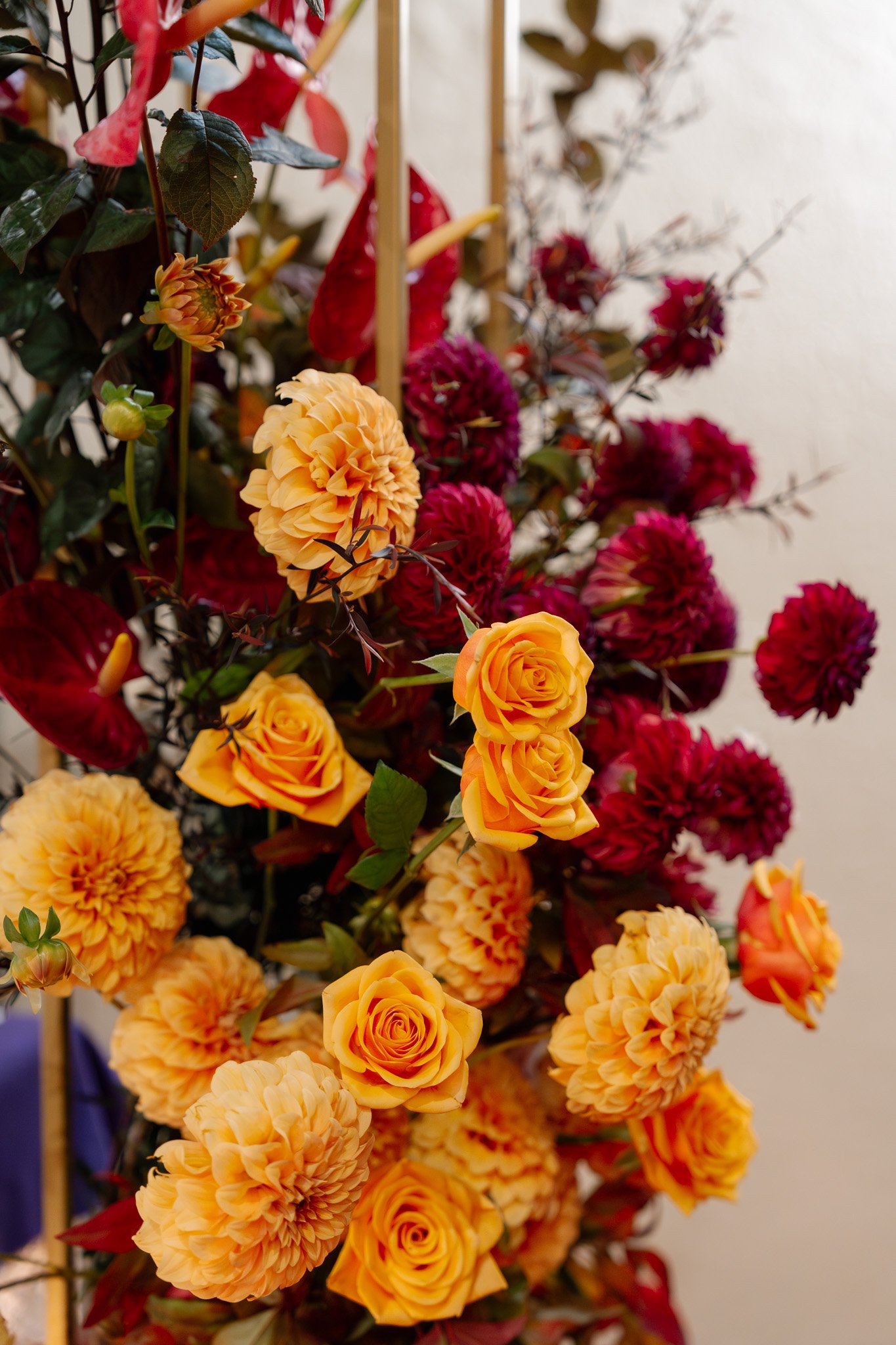 A tall floral arrangement featuring orange roses, peach dahlias, deep red dahlias, and red anthuriums, with a gold support structure against a neutral background.