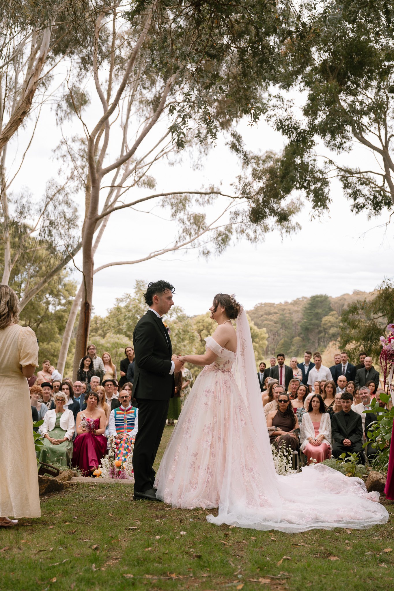 A bride and groom holding hands during an outdoor wedding ceremony under trees, with guests seated and standing in the background.