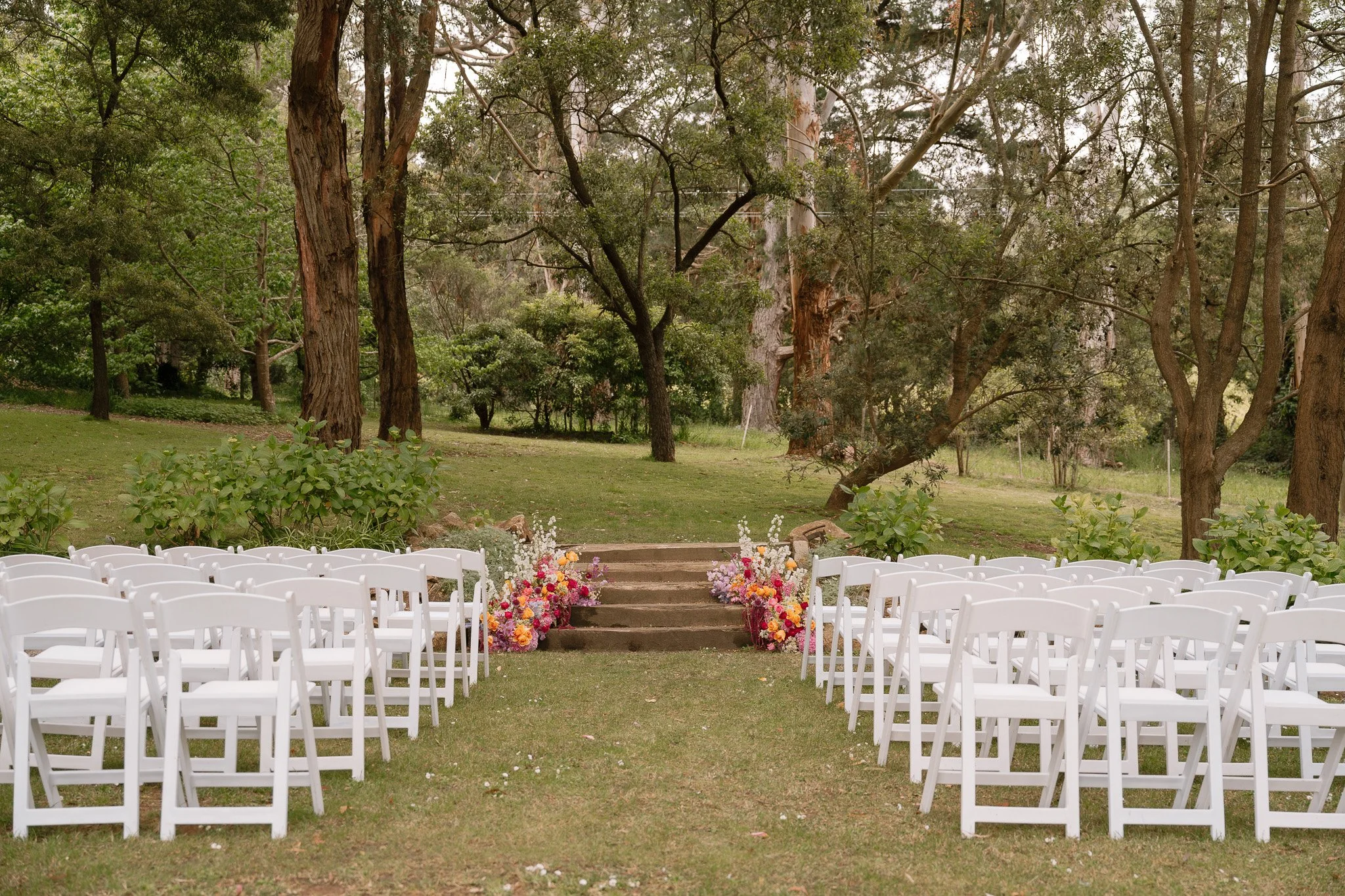 Outdoor wedding ceremony setup with white folding chairs arranged in rows on either side of a grass aisle, decorated with colorful flowers at the steps leading to a wooded area with tall trees and lush greenery in the background.