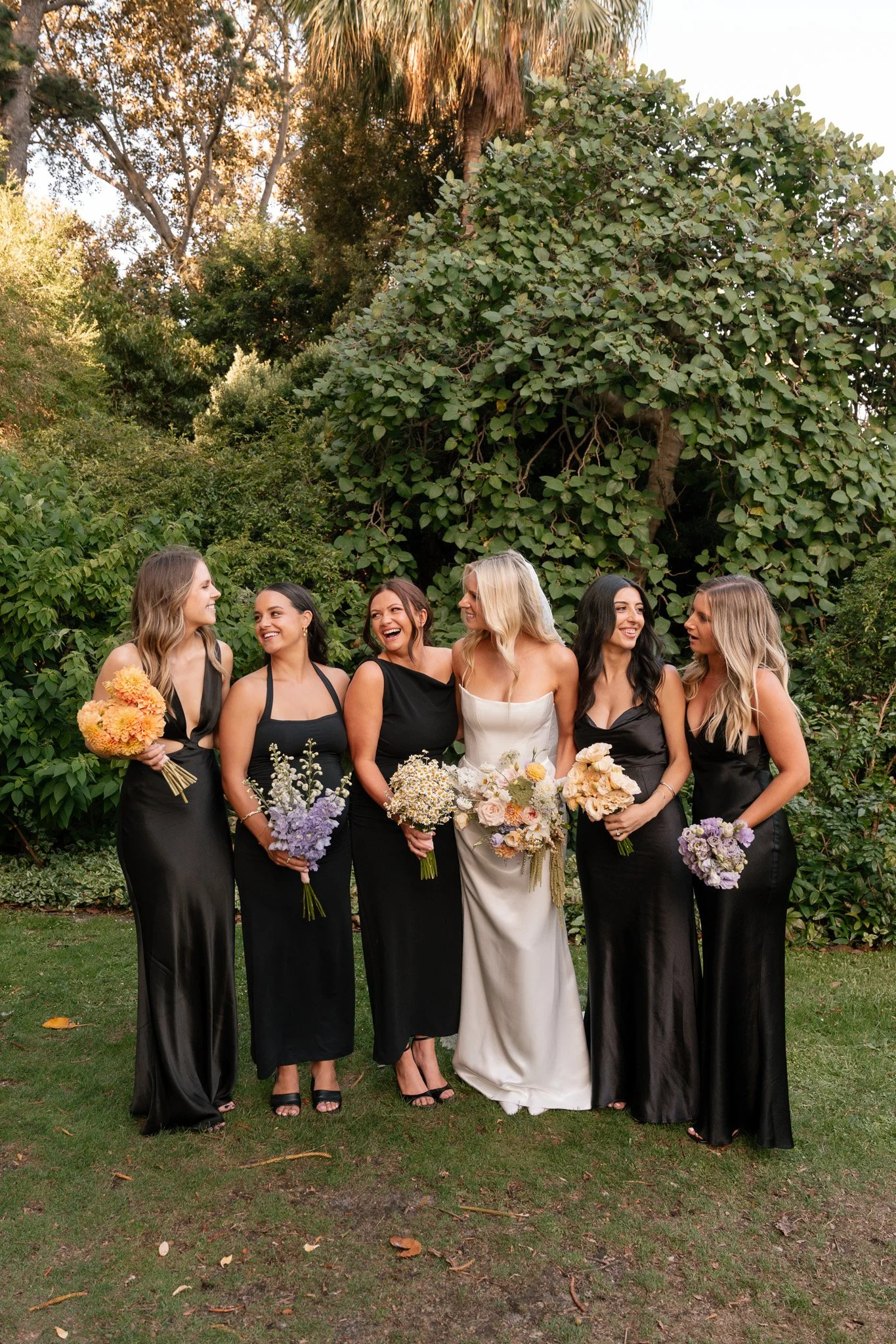 A bride and five bridesmaids standing outdoors in front of green bushes and trees, all holding bouquets of flowers, with the bride in a white wedding dress and the bridesmaids in black dresses, smiling and laughing together.
