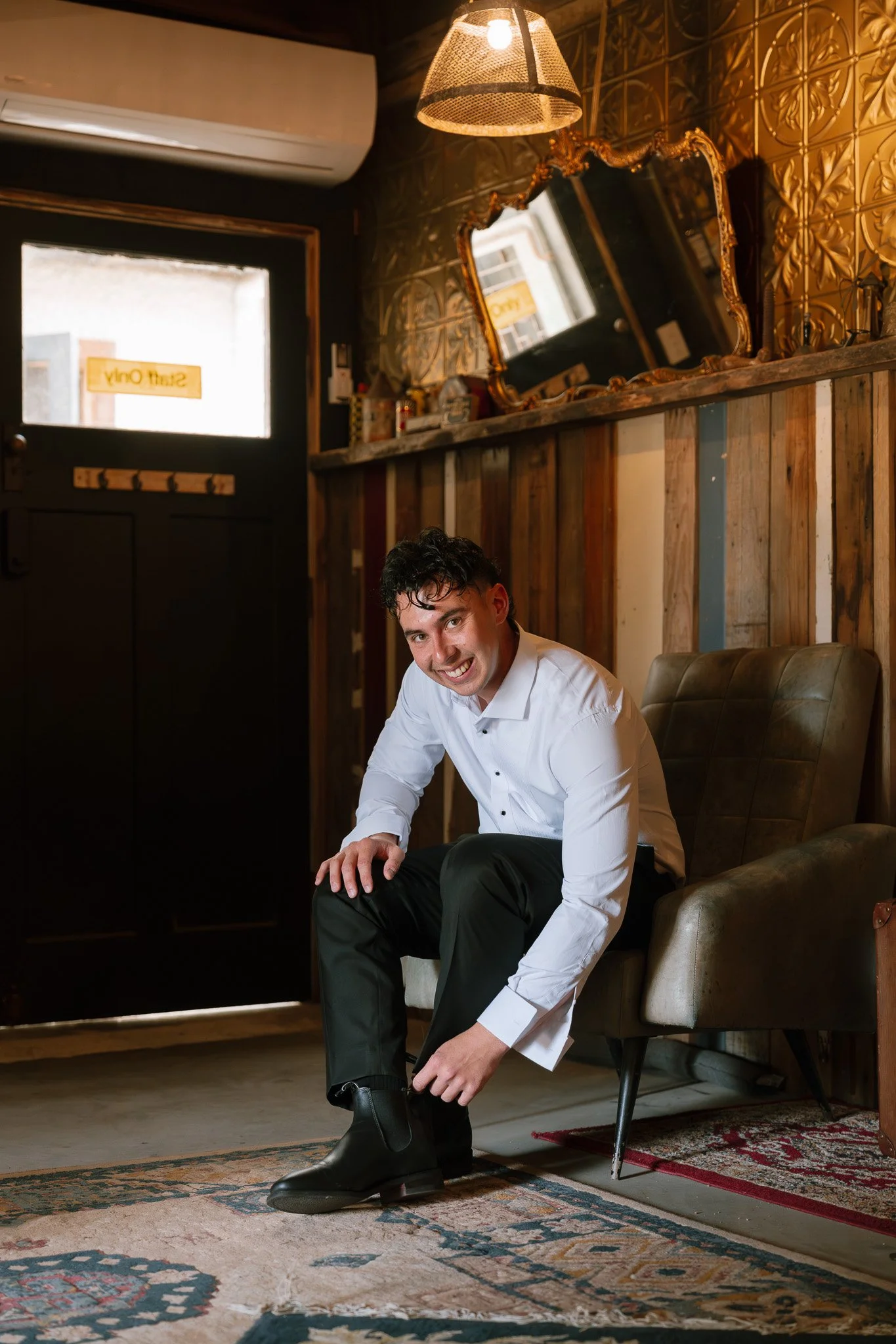 A young man in a white shirt is sitting on a vintage leather armchair, putting on black boots, in a cozy cafe or restaurant with wooden wall paneling and a decorative mirror.