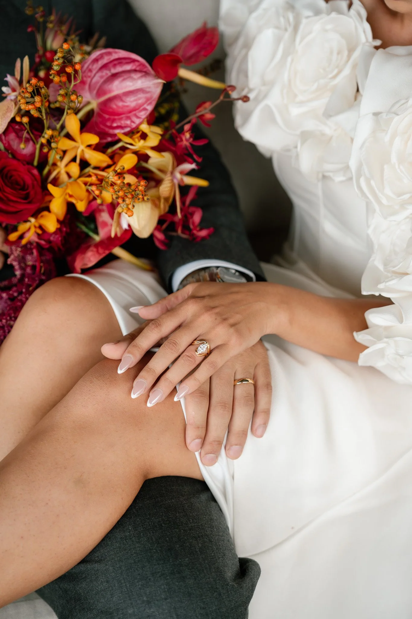 A bride and groom sitting close together, with the bride resting her hand with a wedding ring on her finger on the groom's knee. The groom is holding a bouquet of colorful flowers, including pink, yellow, and red blooms, and is wearing a tuxedo with 