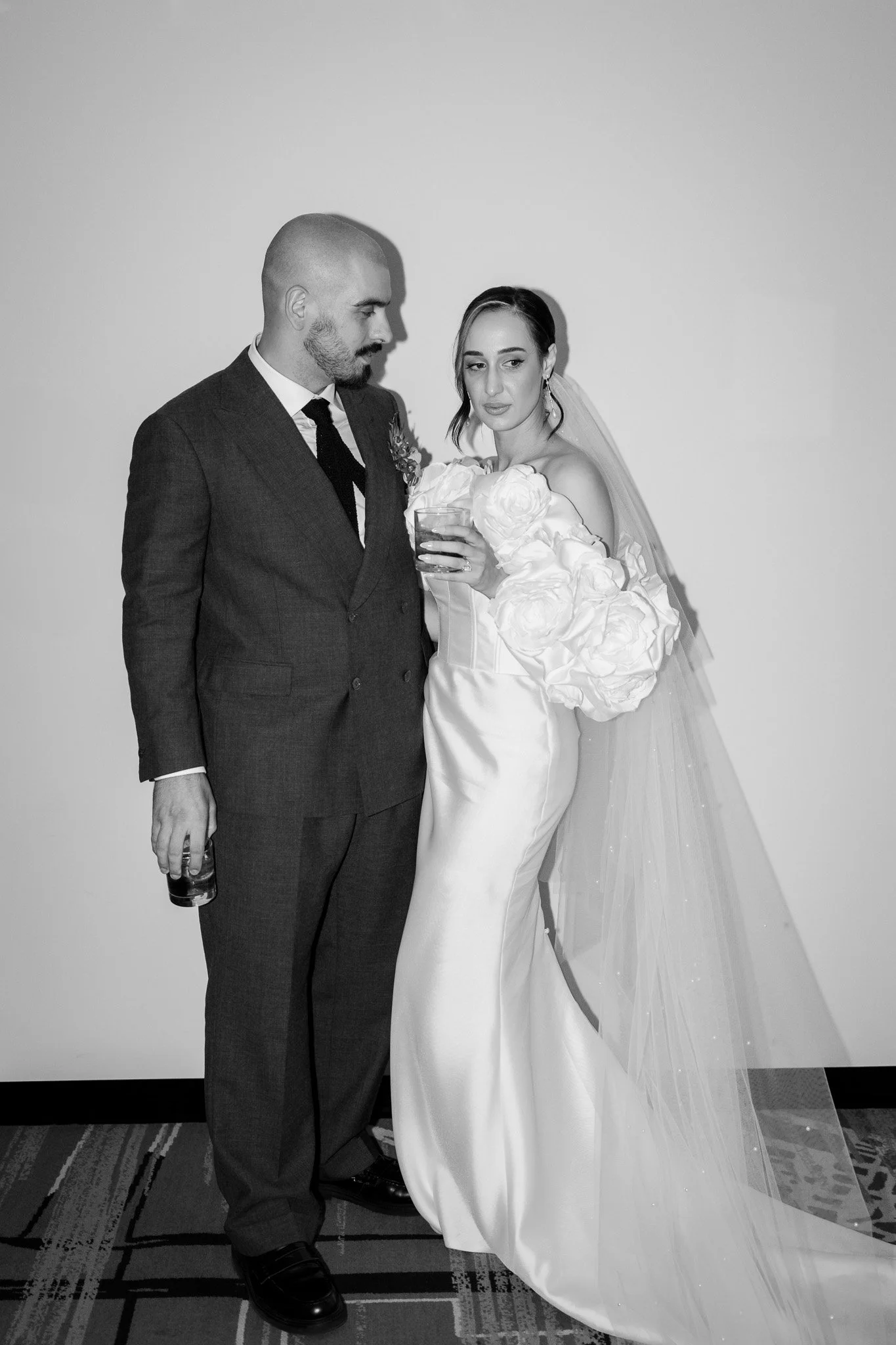 A black and white photo of a bride and groom at their wedding, standing indoors on a carpeted floor against a plain wall. The groom is wearing a dark suit and tie, holding a drink, and looking at the bride. The bride is wearing a long, fitted wedding