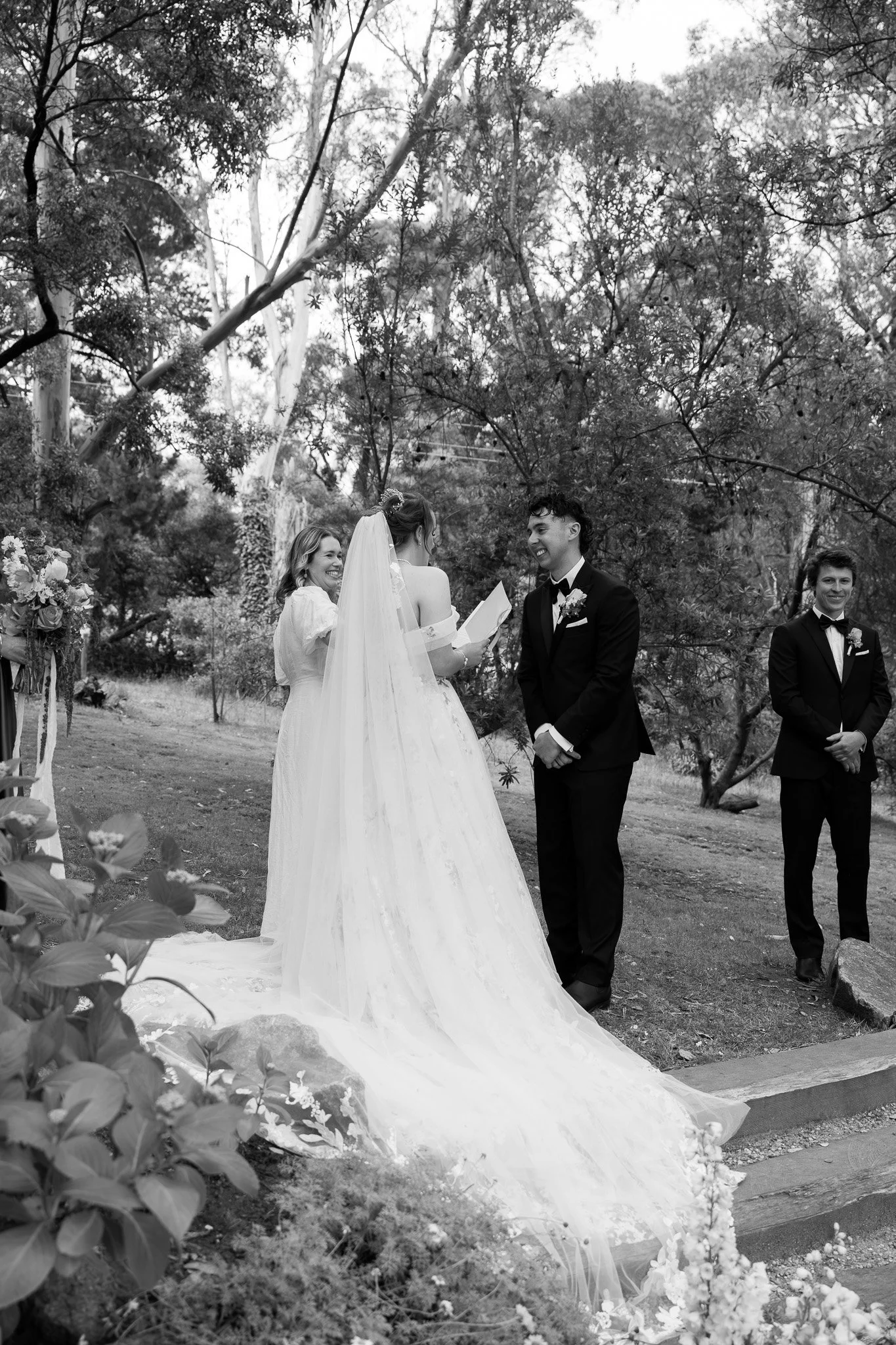 Black and white photo of a wedding ceremony outdoors, showing a bride and groom standing at the altar, with a female officiant and a groomsman. The bride is wearing a long wedding gown and veil, and the groom is in a tuxedo. Trees and flowers surroun