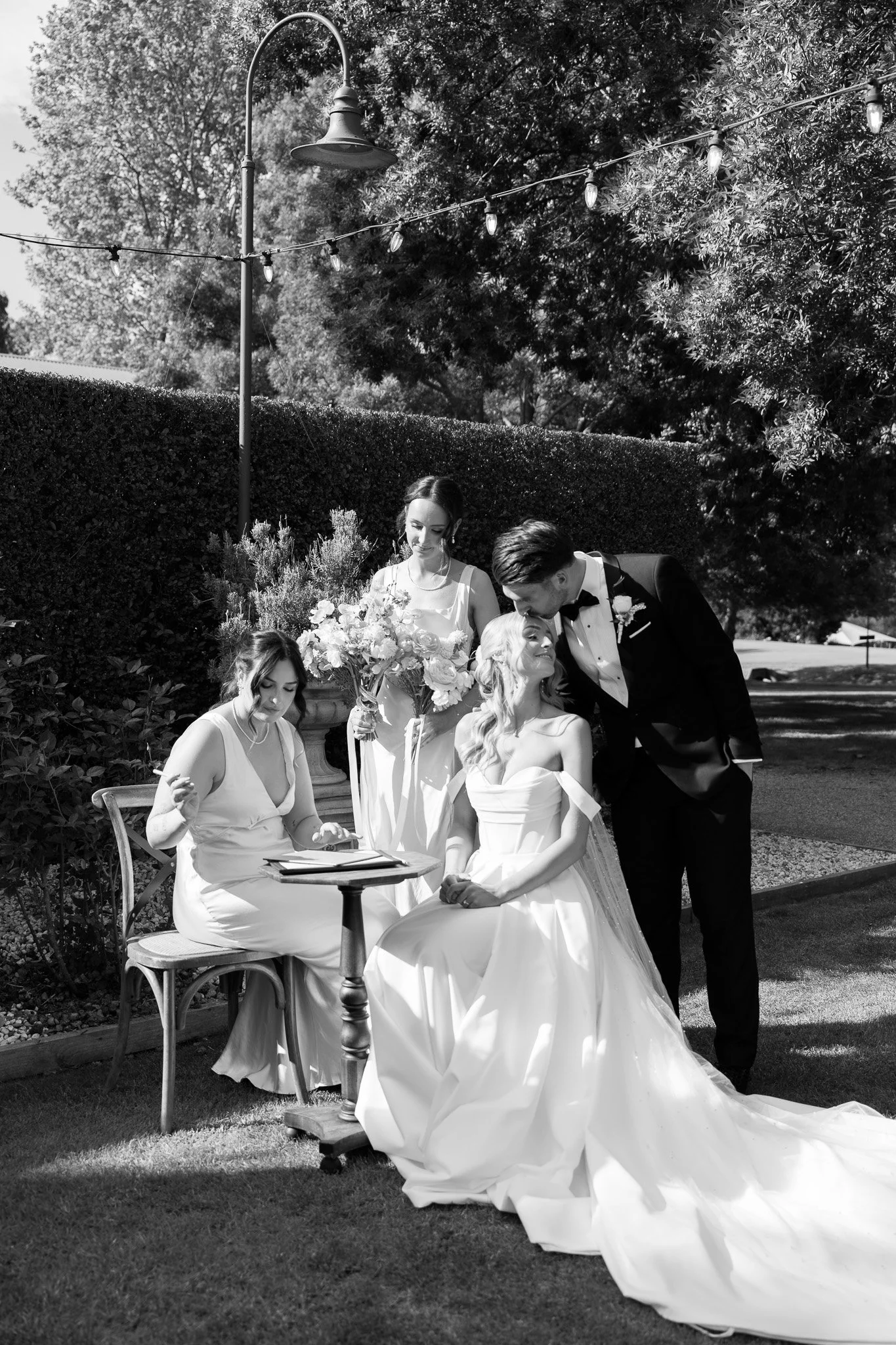 Black and white wedding scene outdoors with bride, groom, and two women, one arranging flowers and one signing a document.