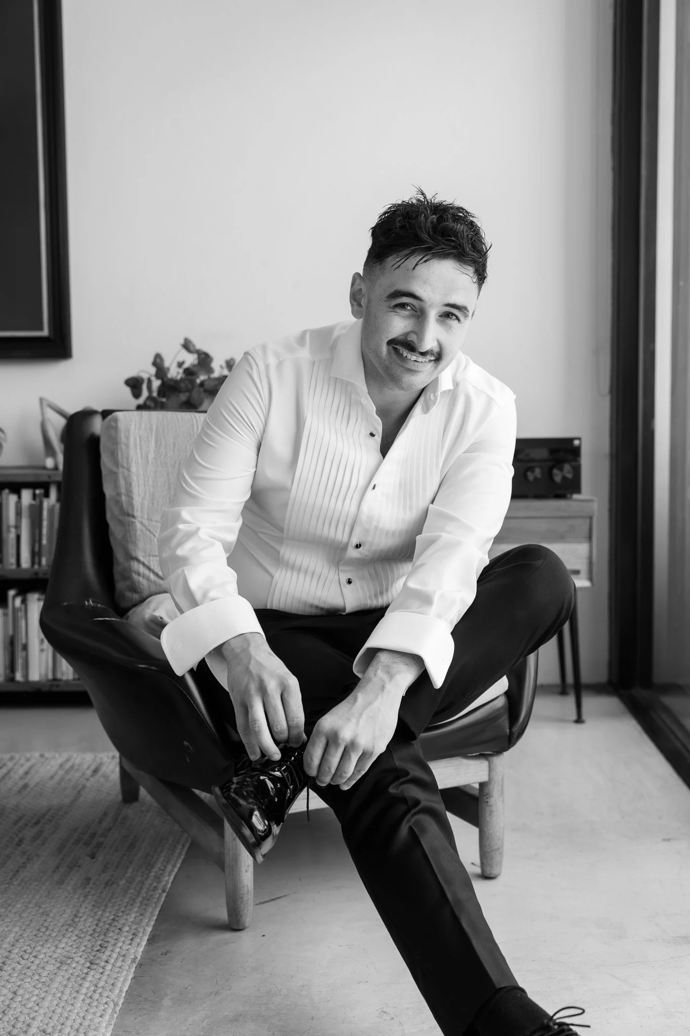 A smiling man with a mustache, wearing a white dress shirt, black pants, and shiny black shoes, is sitting on a chair near a window, tying his shoelace in a modern living room.