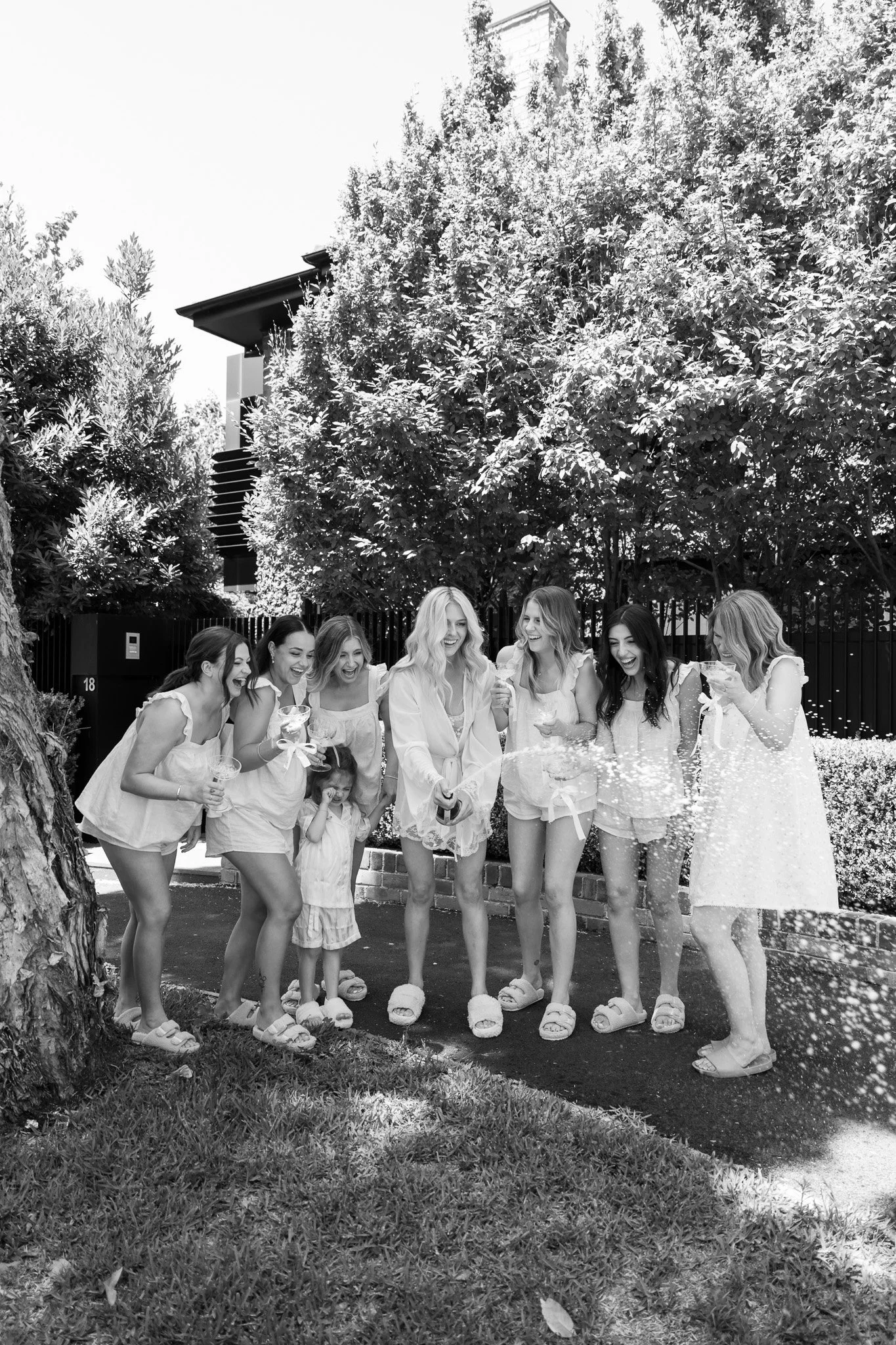 A group of women and girls celebrating outdoors, opening a bottle of champagne with sparklers and party poppers, smiling and wearing casual summer clothing.