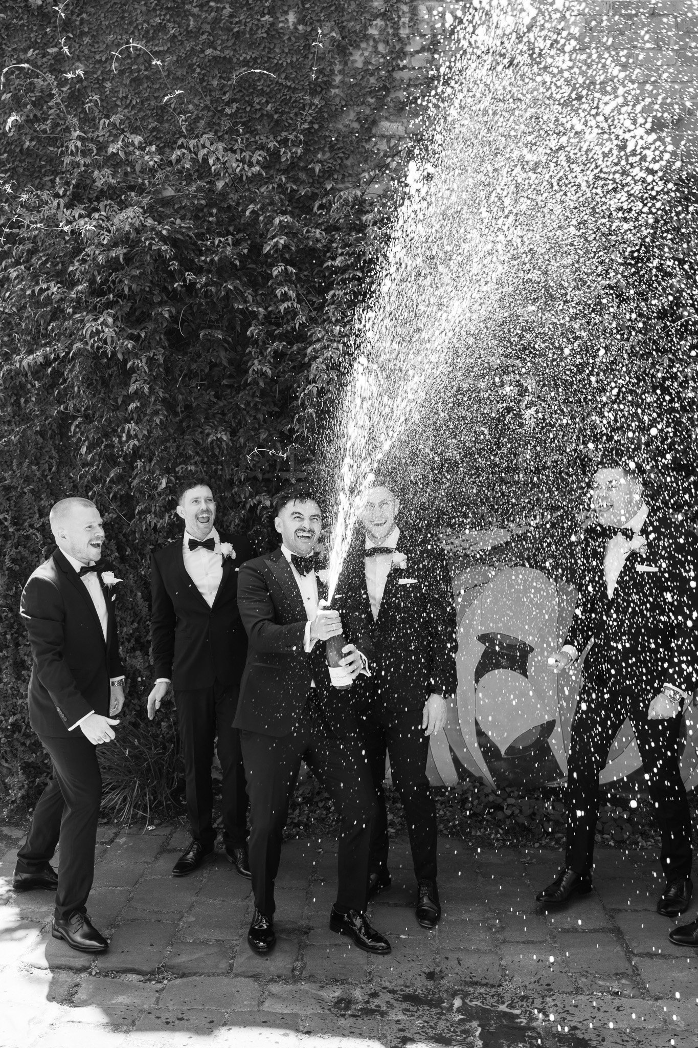 Group of men in tuxedos celebrating with champagne, spraying it into the air, outdoors near a wall with decorative art.