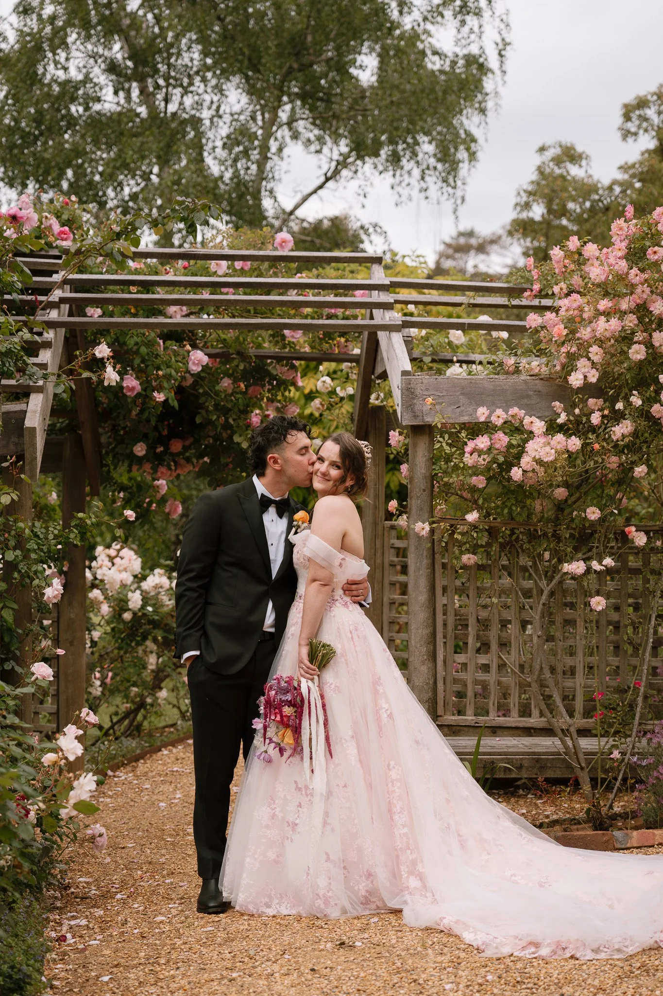 A wedding couple standing on a garden path surrounded by blooming pink roses, with the groom kissing the bride on the cheek.