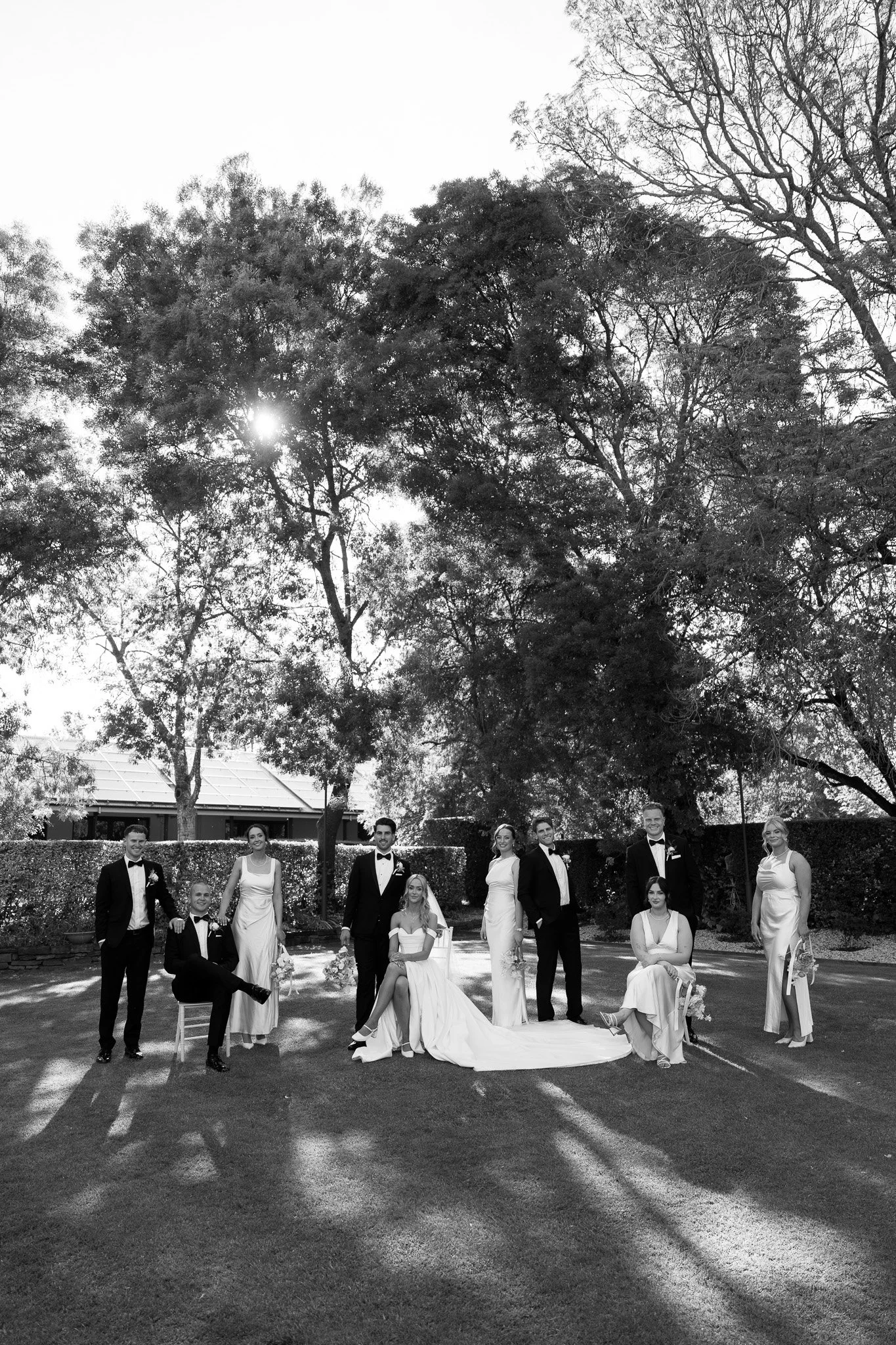 A black and white wedding photo featuring a bride in a wedding gown and veil, surrounded by bridesmaids in matching dresses and groomsmen in tuxedos, outdoors on a lawn with trees and a building in the background.