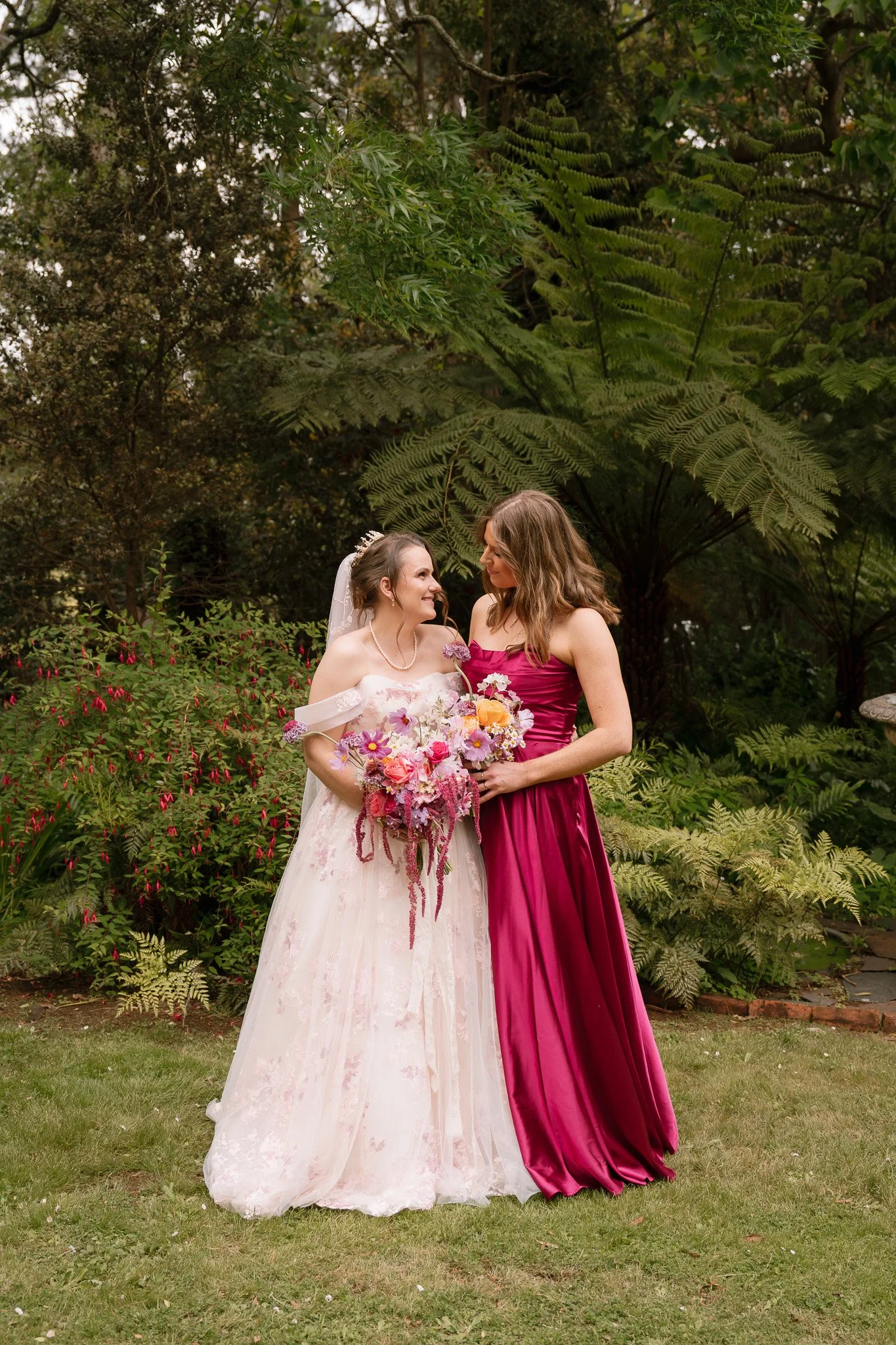 A bride in a white wedding gown holding a colorful bouquet standing next to a woman in a deep pink dress. They are outdoors in a garden with lush green foliage.