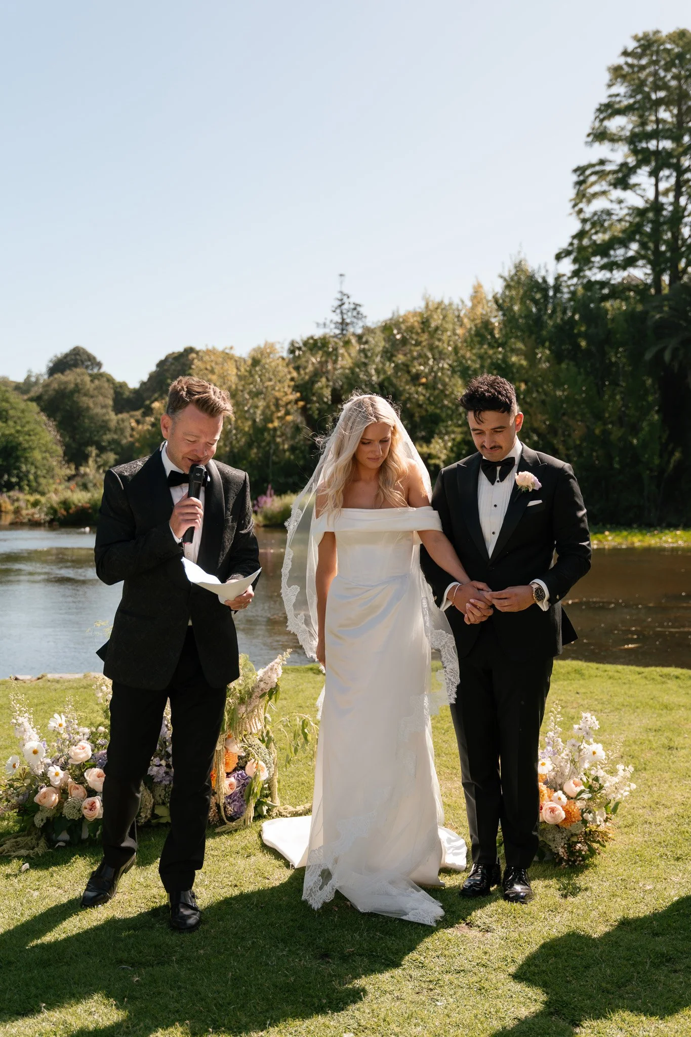 A wedding ceremony outdoors by a lake with three people: a officiant reading from paper, a bride in a white gown with veil, and a groom in a black tuxedo holding bride's hand, surrounded by floral arrangements.