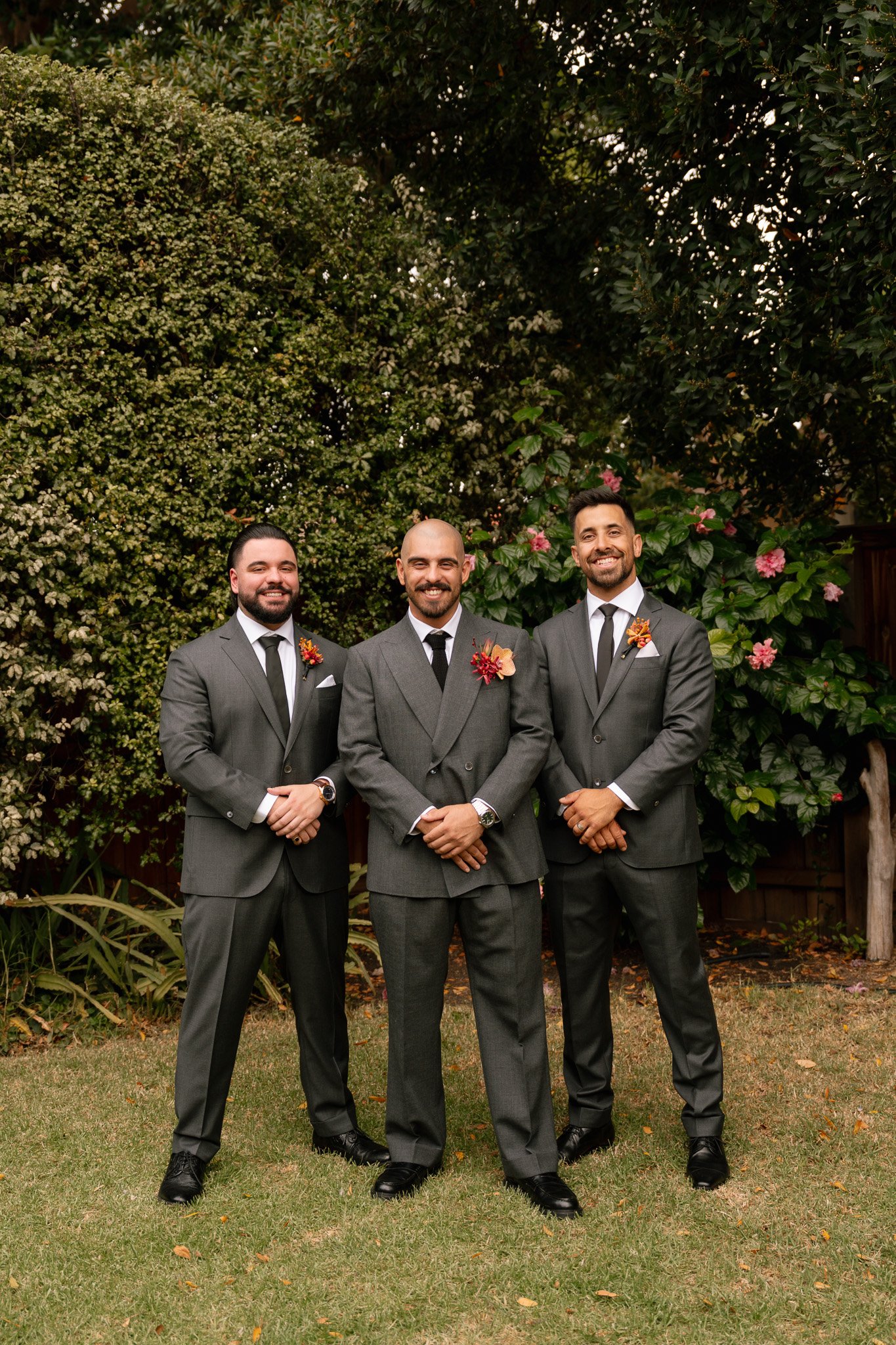 Three men in dark gray suits standing outdoors on grass, smiling, with greenery and flowering bushes in the background.