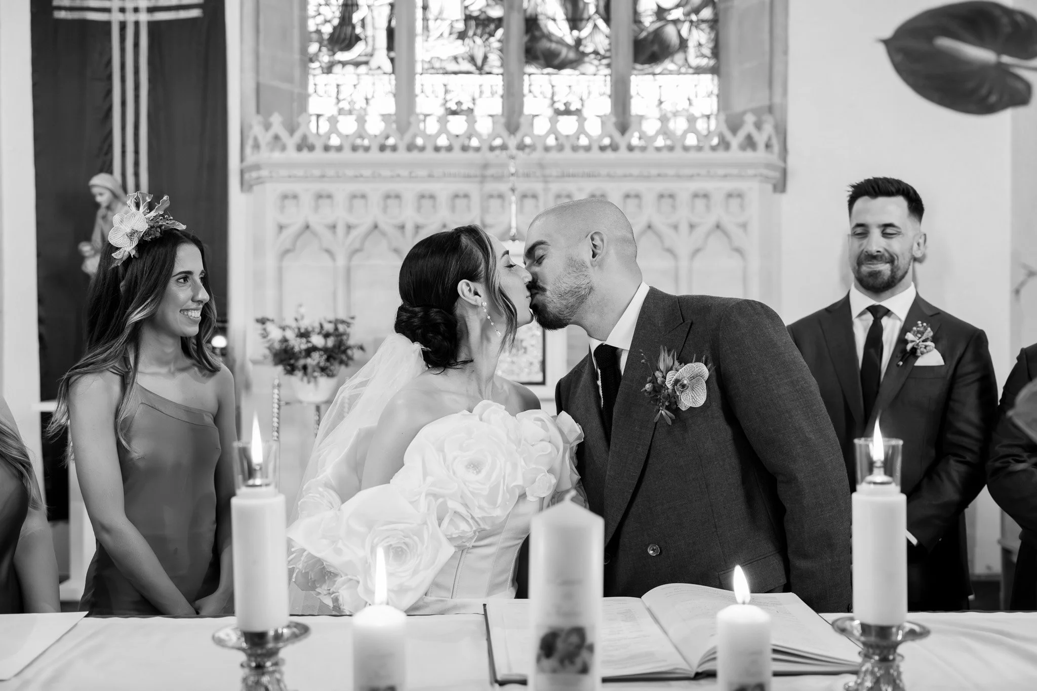 A black and white photo of a wedding ceremony where a bride and groom are kissing. The bride is wearing a wedding dress with floral details and a veil, while the groom is in a suit with a boutonniere. There are bridesmaids and groomsmen standing arou