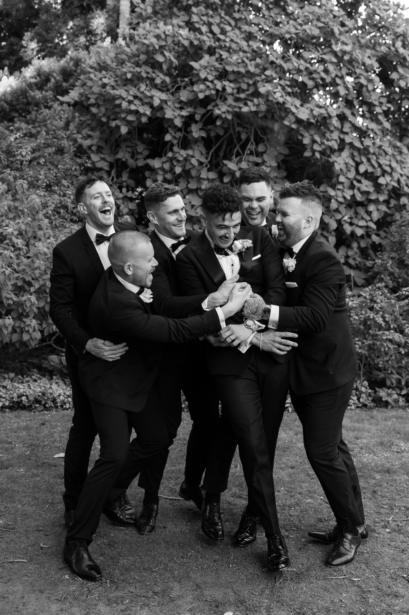 A group of seven men in tuxedos laughing and celebrating outdoors. They are holding hands and hugging each other, standing on grass with large bushes in the background.