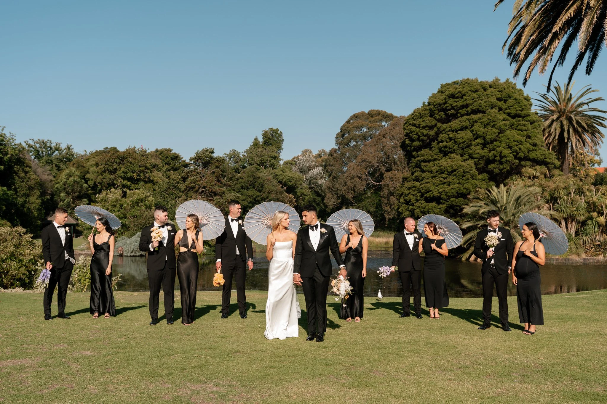 A wedding party on grass near a lake, with trees and blue sky in the background. The bride and groom walk in front, holding hands, with bridesmaids and groomsmen behind them holding umbrellas.