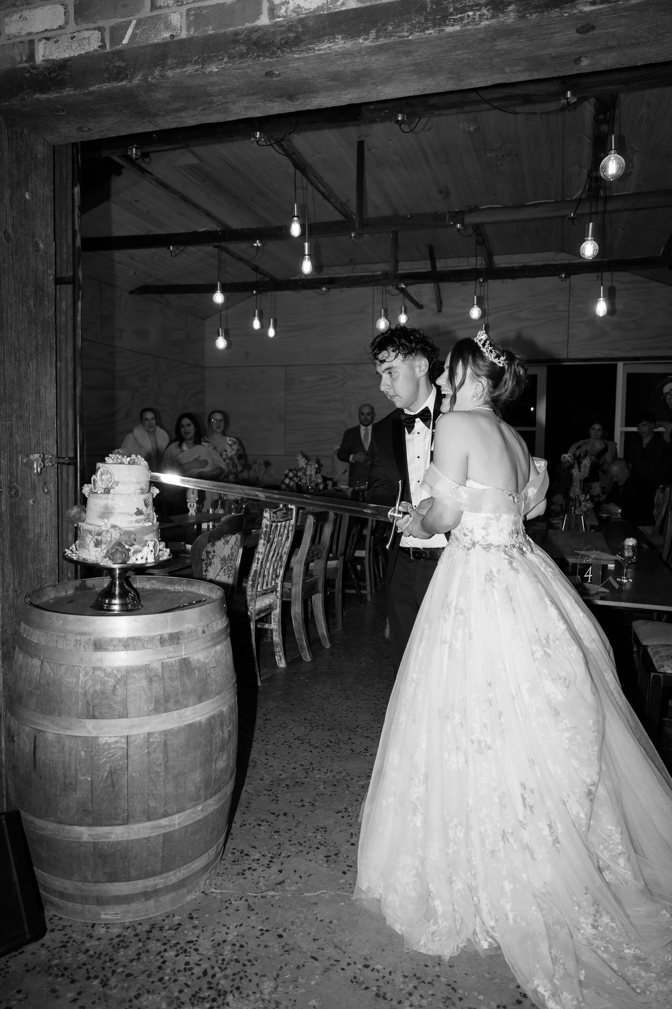 A bride and groom at their wedding reception, lighting a celebratory firework candle on a cake placed on a barrel, with guests watching in the background.