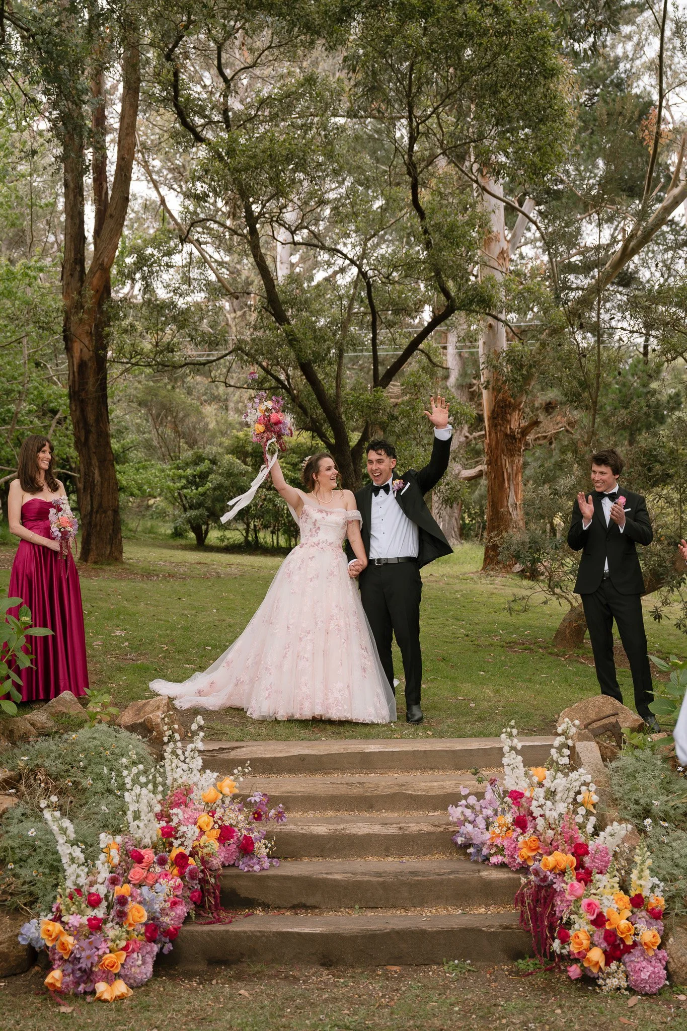 Bride and groom celebrating at outdoor wedding ceremony, surrounded by bridesmaids and groomsmen, colorful flower arrangements on steps, in a wooded area.