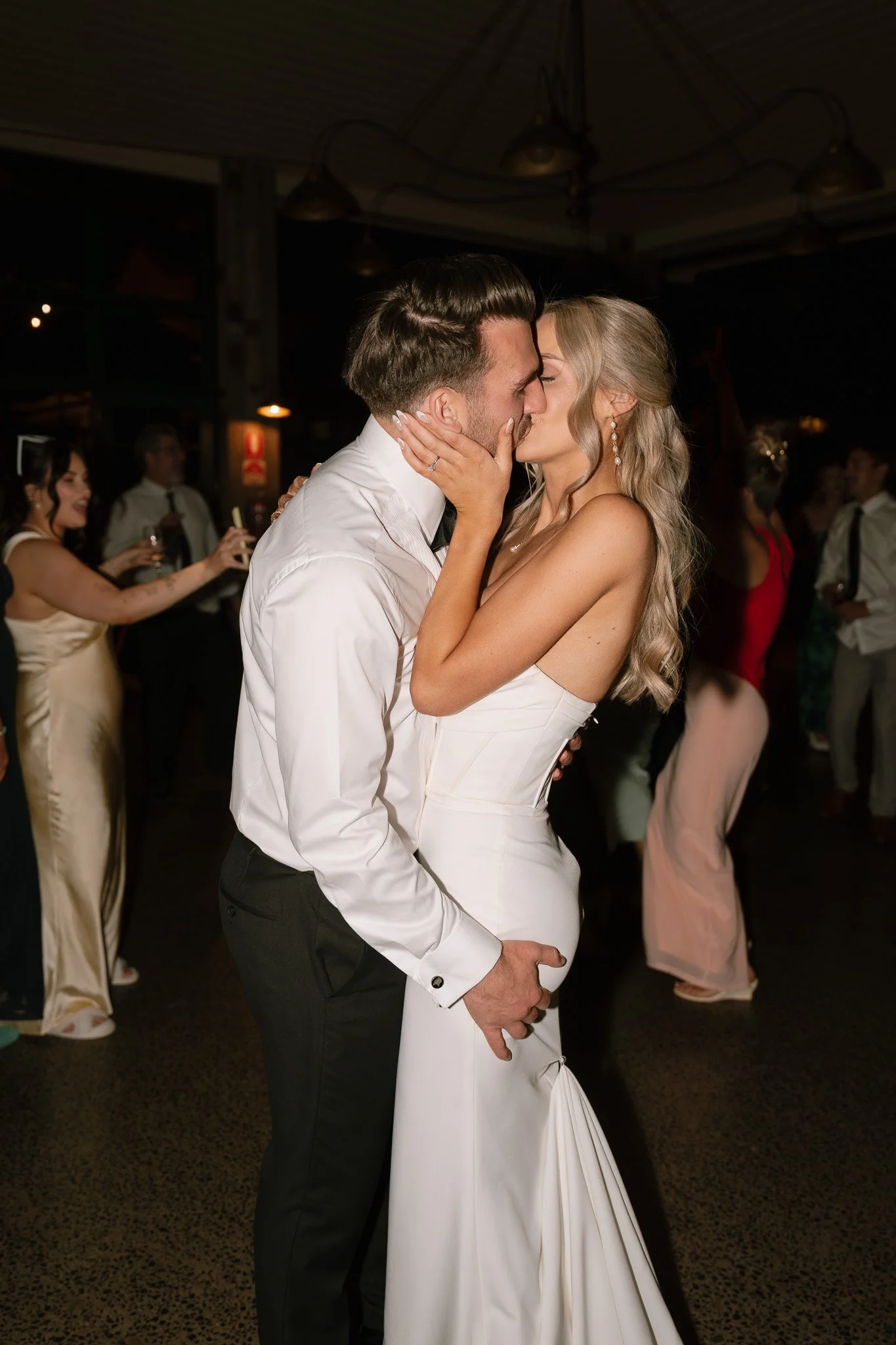 A couple is kissing on the dance floor at a wedding reception, with guests in the background celebrating.