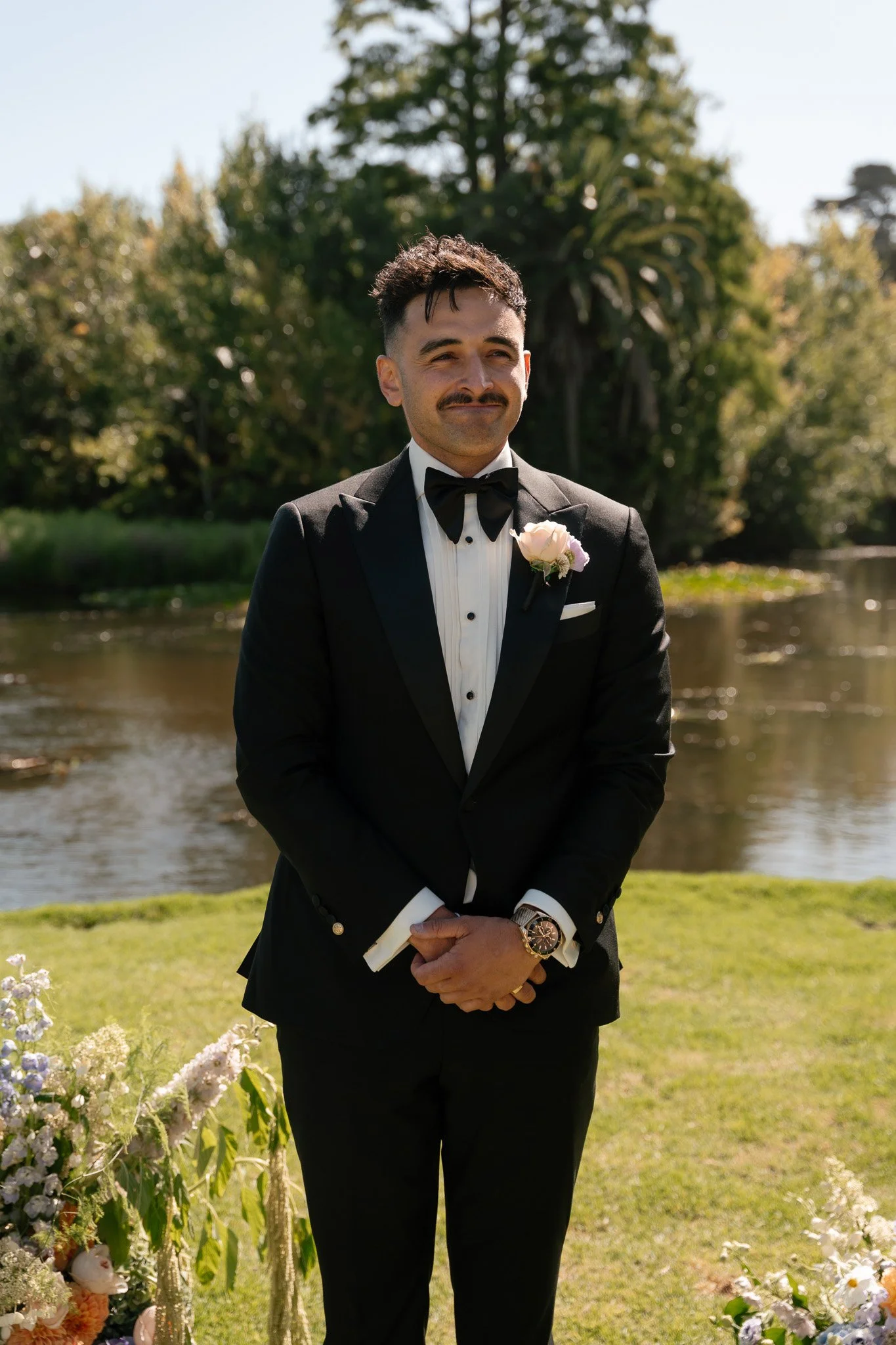 A man dressed in a black tuxedo with a bow tie and a boutonniere, standing outdoors by a river, with trees in the background, and floral decorations in the foreground.
