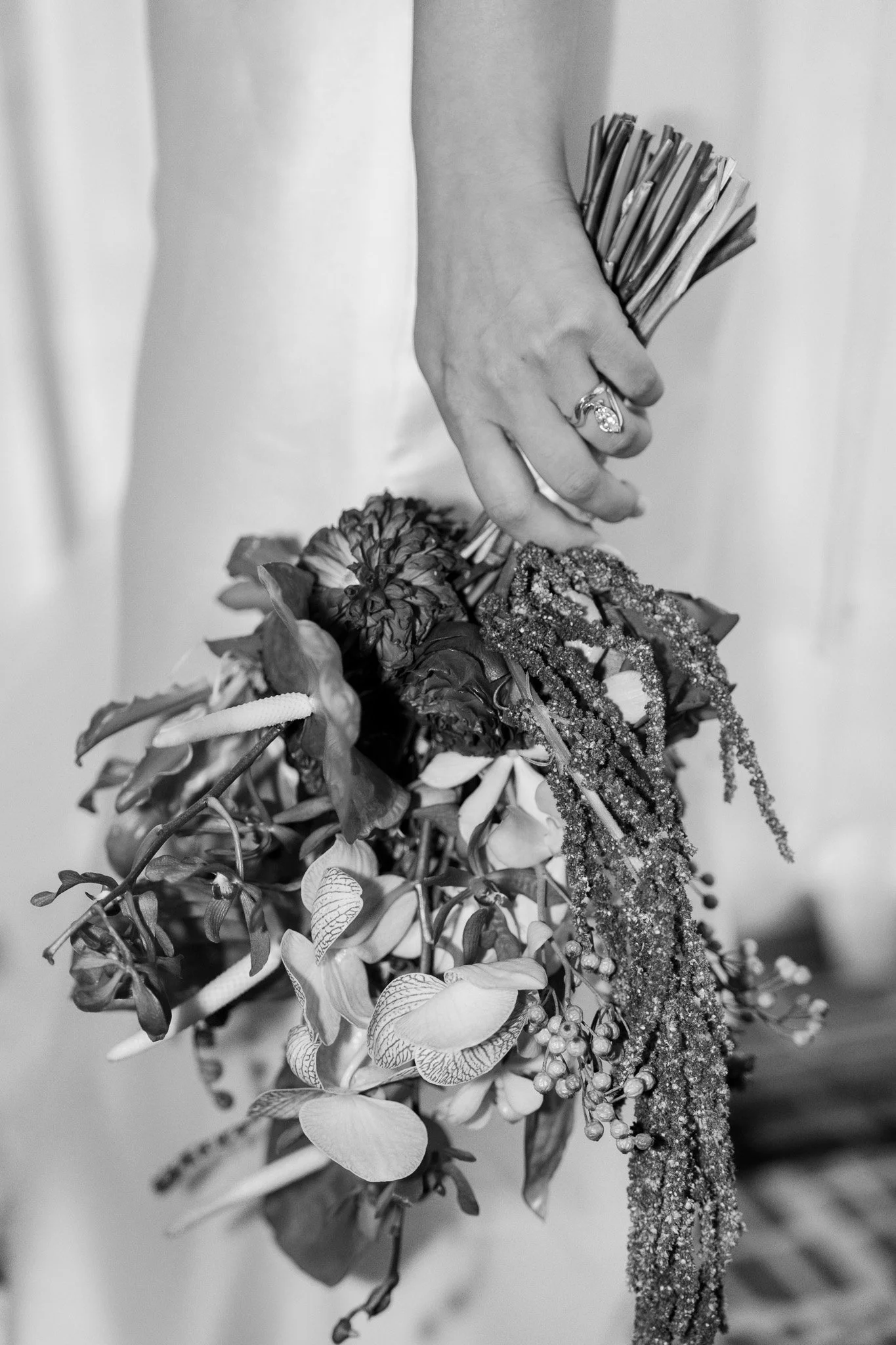 A close-up of a woman's hand holding a bouquet of flowers, including orchids and other blooms, with a wedding ring visible on her finger, in black and white.