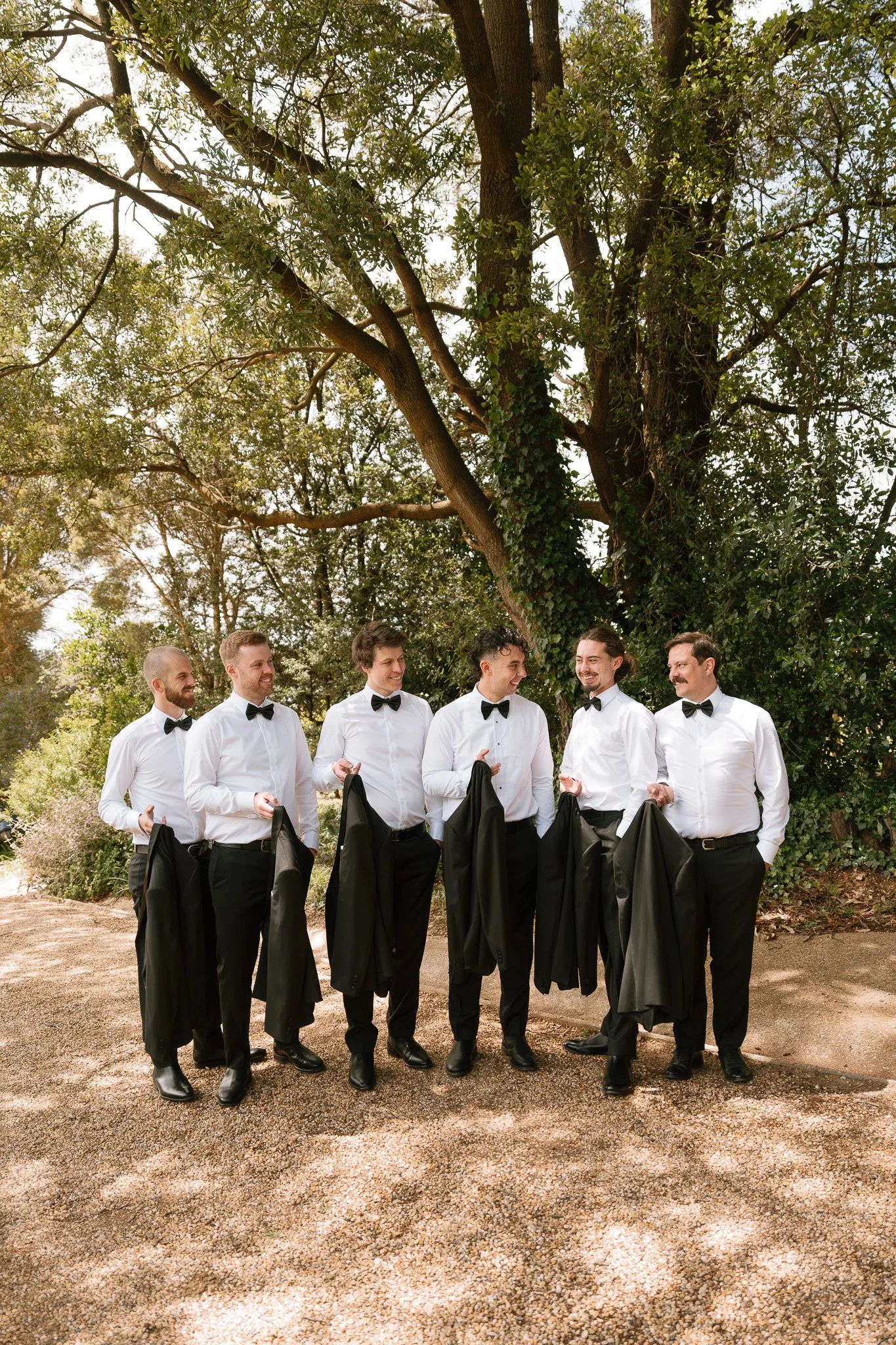 Six men dressed in white shirts, black bow ties, and black pants holding black jackets, standing outdoors under a large tree, smiling and talking.