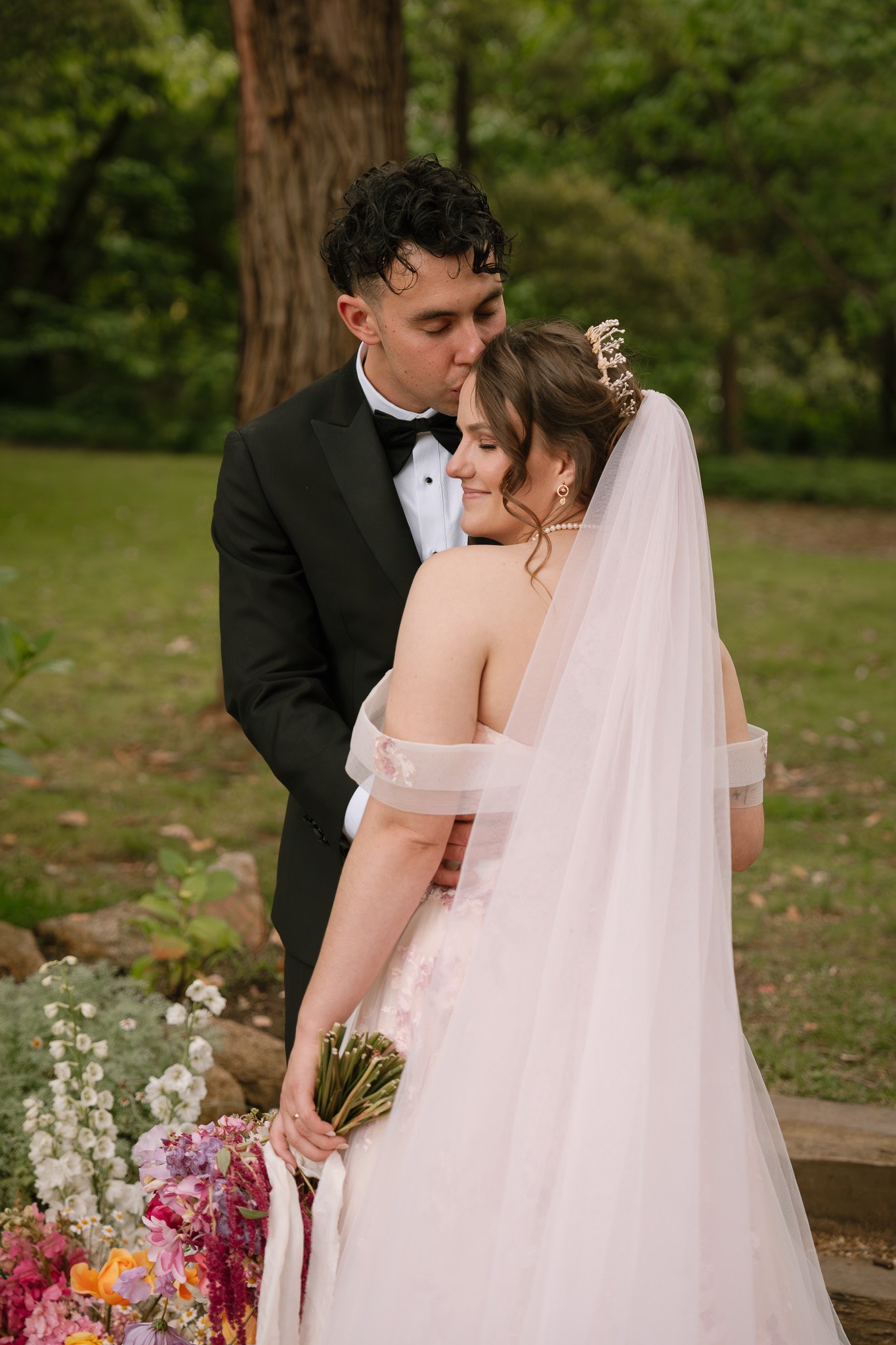 A bride and groom sharing a kiss outdoors, surrounded by flowers and trees.