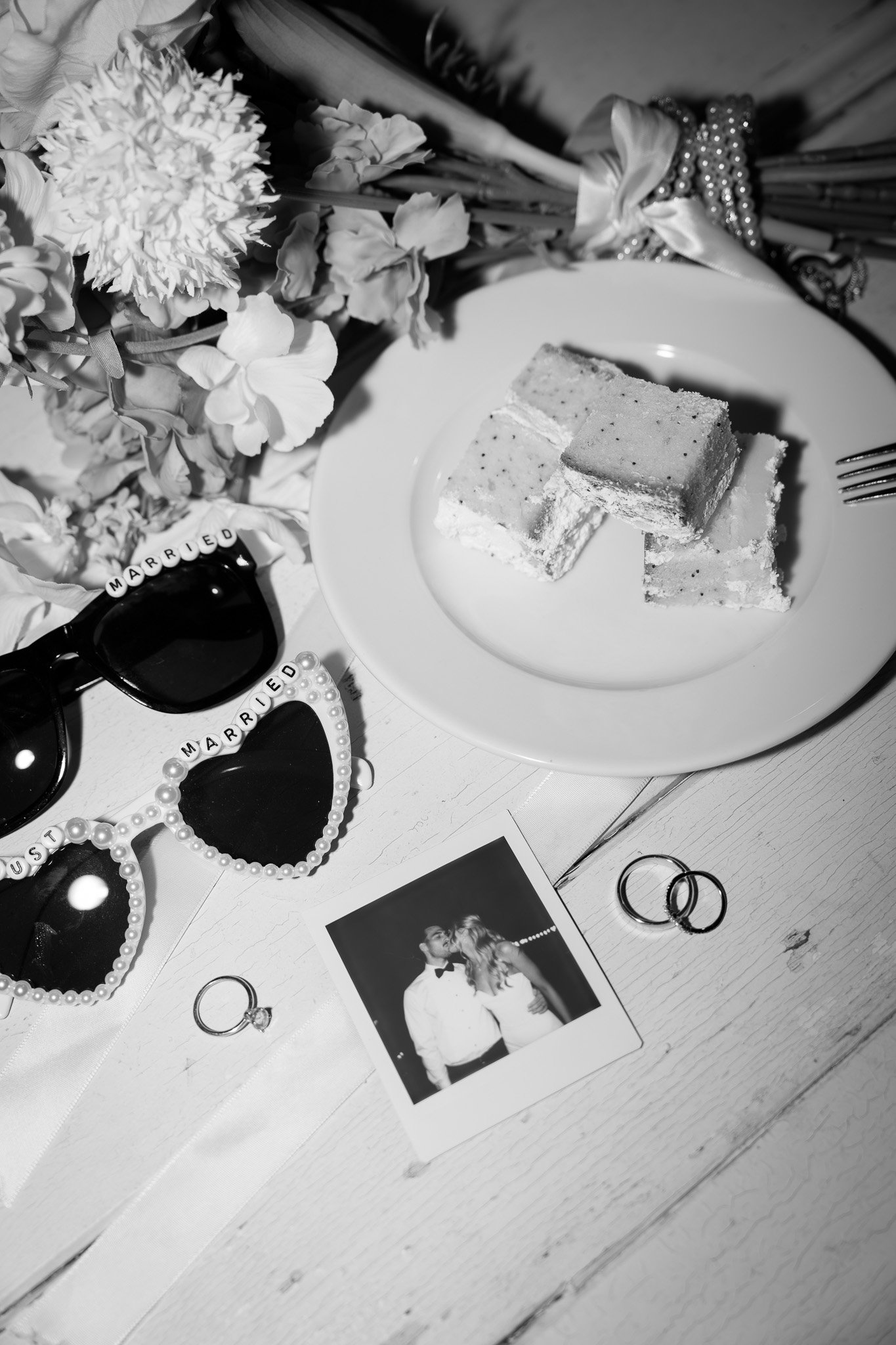 A black-and-white photo of a wedding anniversary celebration table with a bouquet of flowers, sunglasses with "Married" spelled out on beads, wedding rings, a photo of a couple, a napkin, and a plate with layered cake.