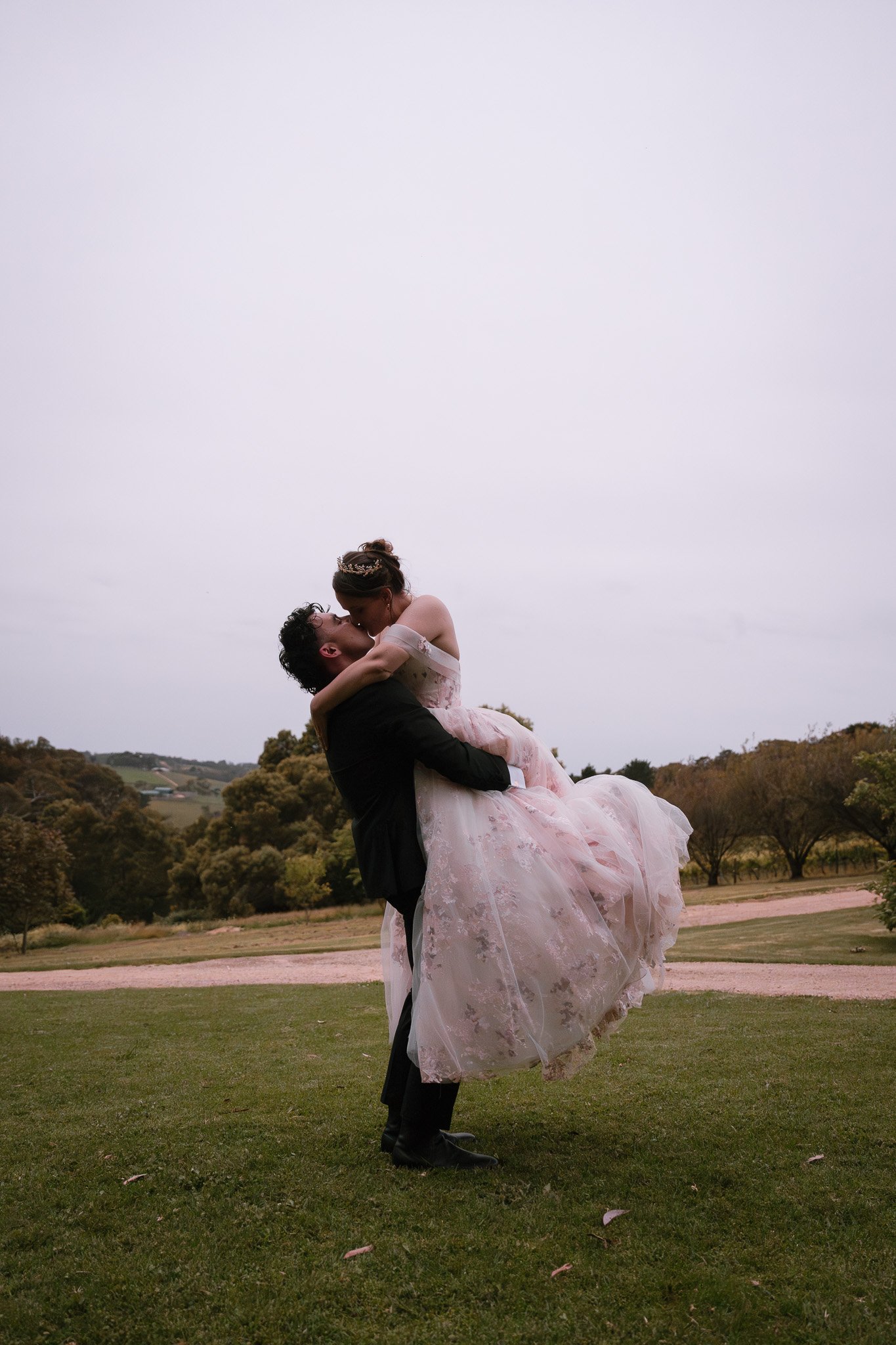 A couple in wedding attire sharing a romantic kiss outdoors. The man is lifting the woman, who is wearing a pink lace wedding dress, in a grassy area with trees and hills in the background under a cloudy sky.