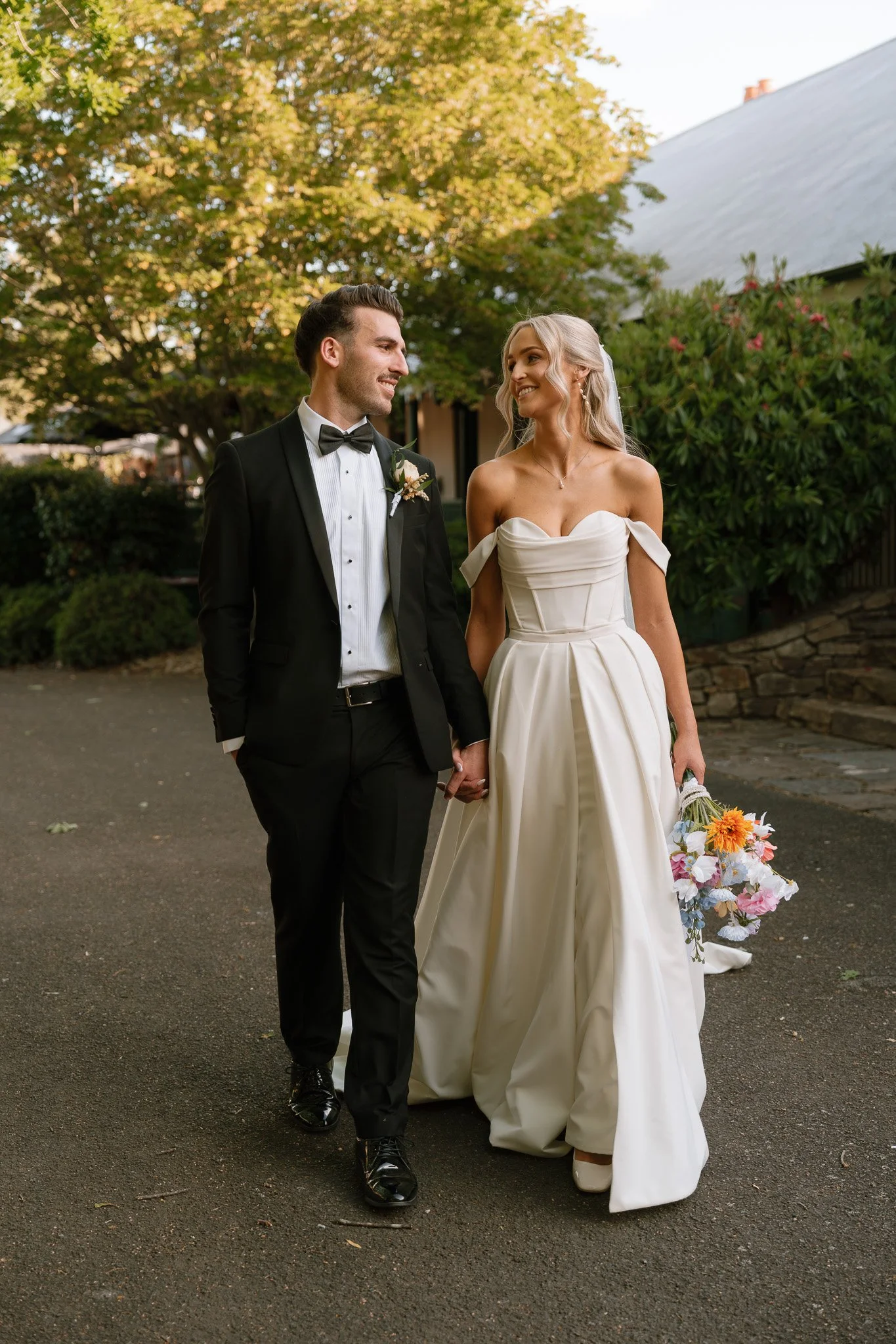 A bride and groom holding hands and walking outdoors, smiling at each other. The bride is wearing a white off-shoulder wedding gown and holding a colorful bouquet, while the groom is dressed in a black tuxedo with a bow tie. There are trees and a bui