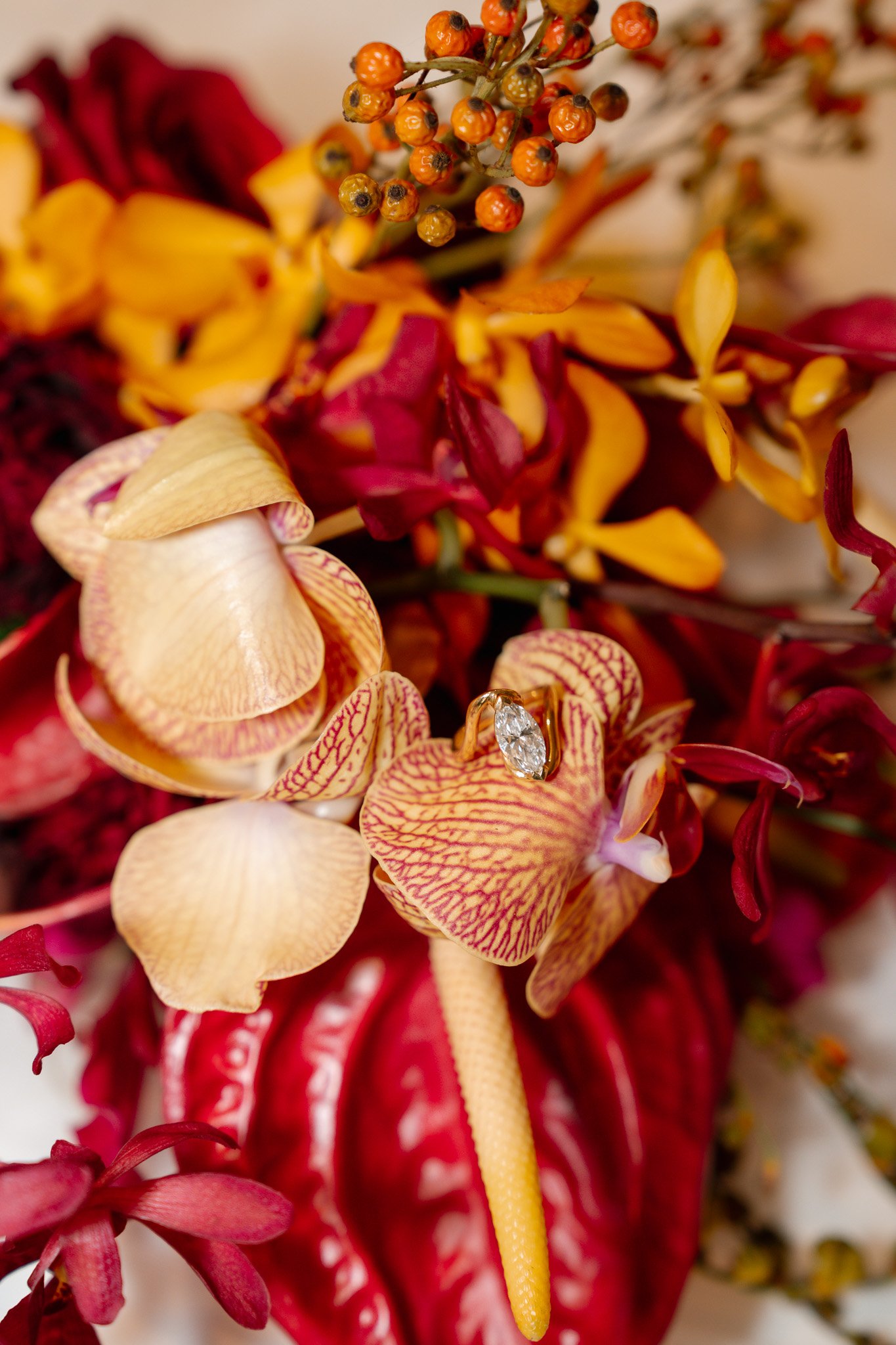 Close-up of a floral arrangement with orchids, colorful leaves, and a diamond ring resting on one of the orchids.