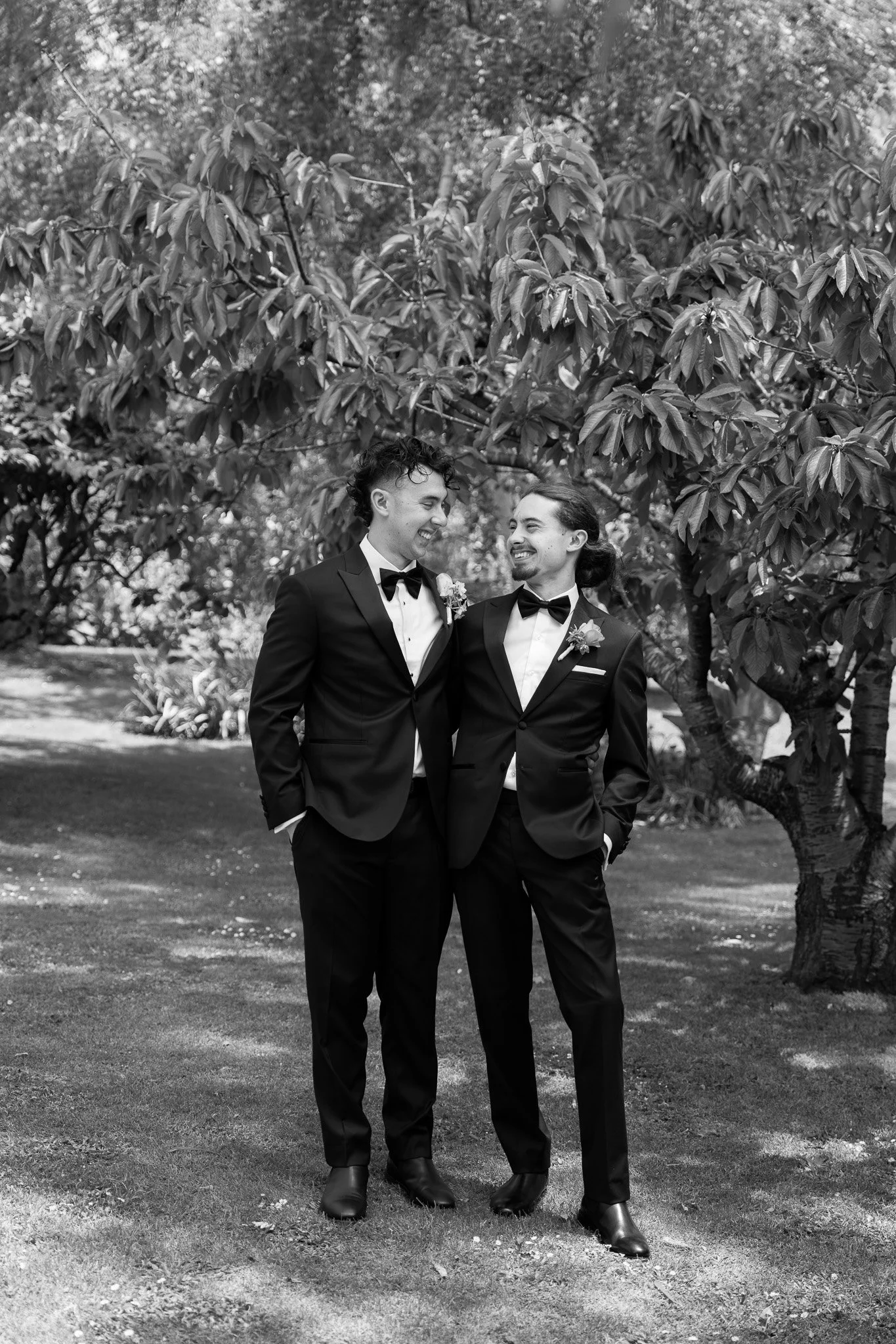 Two men in tuxedos standing close together outdoors, smiling and looking at each other during a wedding or formal occasion, with trees and grass in the background.