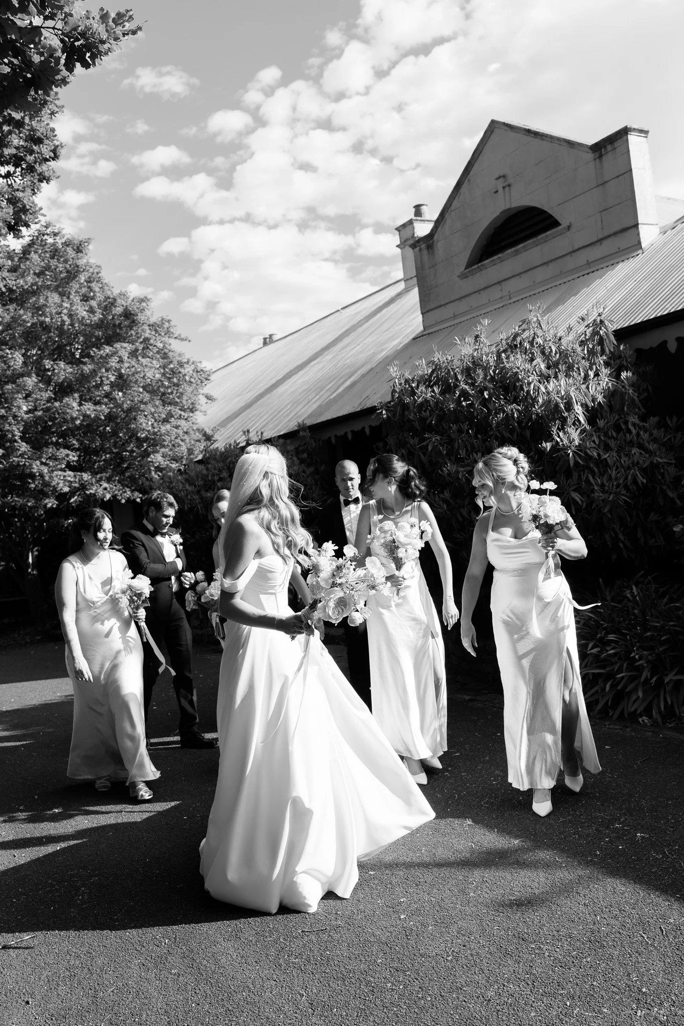 A black and white photo of a bride and bridesmaids walking outdoors, holding bouquets, near a rustic building and trees.