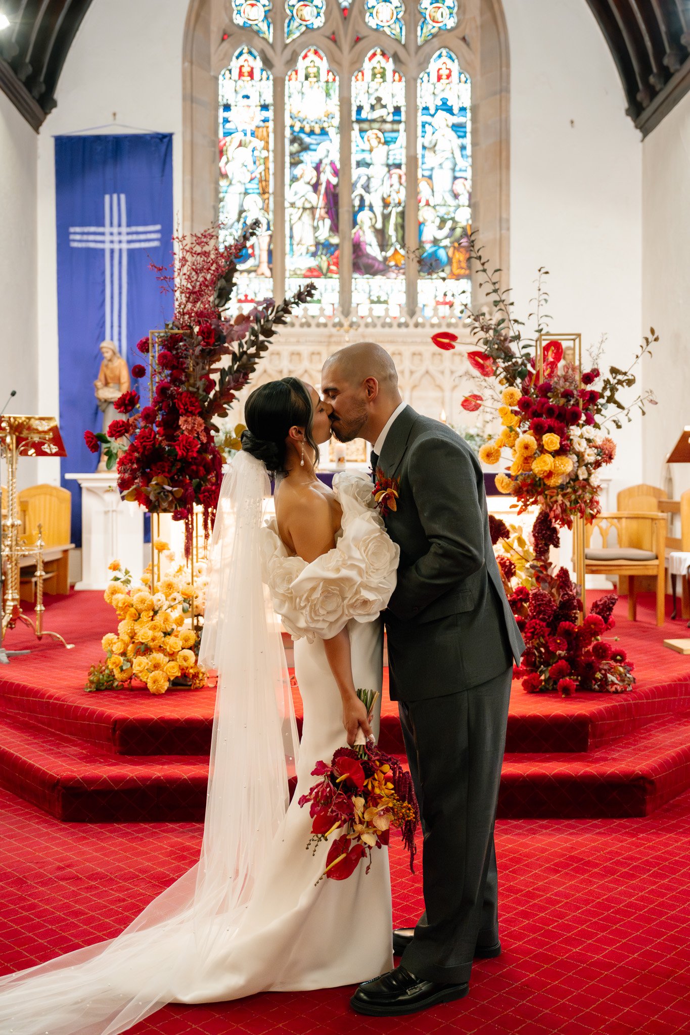 A bride and groom sharing a kiss during their wedding ceremony inside a church with stained glass windows and colorful floral arrangements.